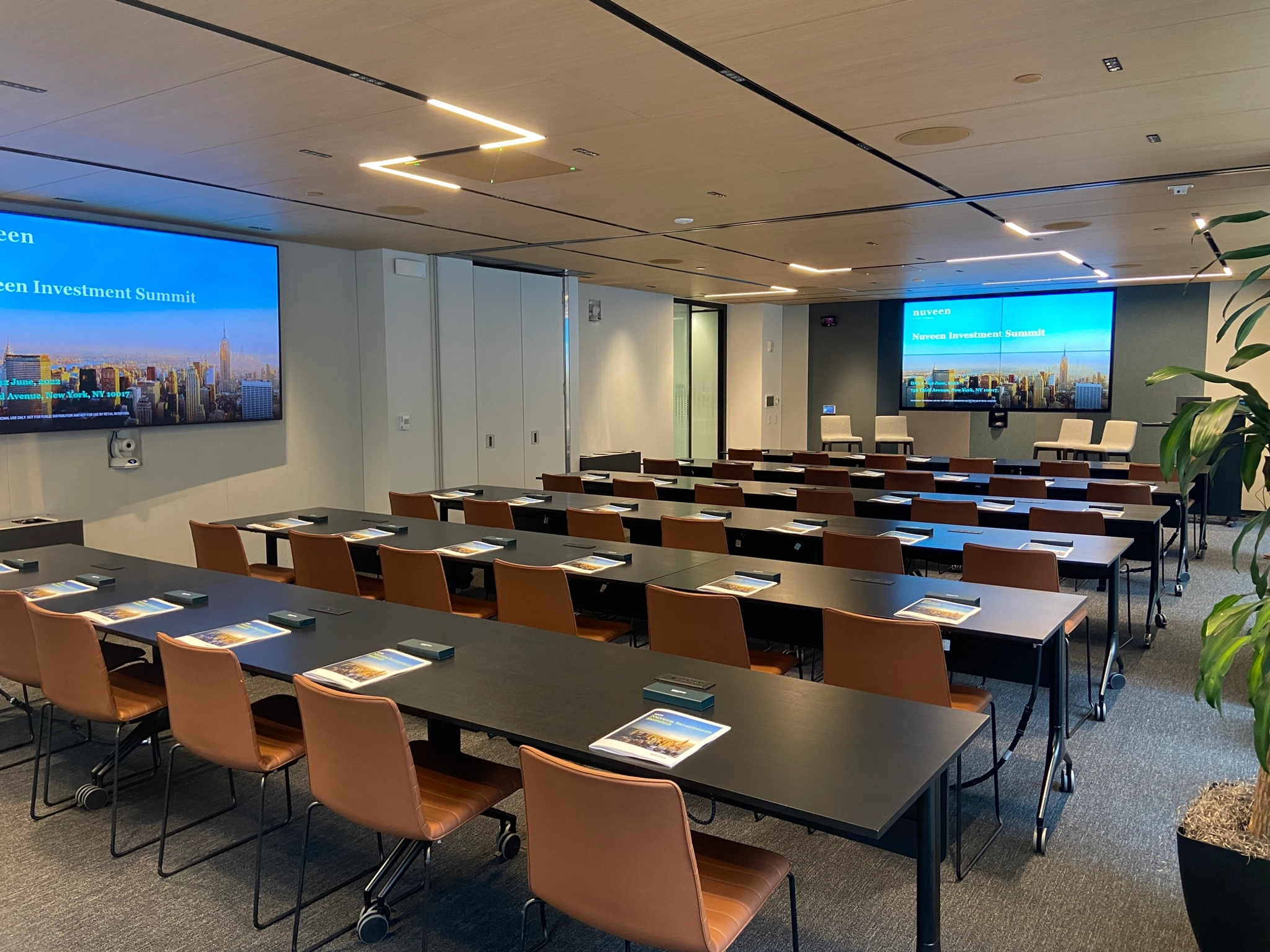 A modern meeting room in New York, featuring rows of tables and chairs, each set with notebooks and water bottles. Two large screens display "Morgan Investment Summit," while plants adorn the back of this coworking space.