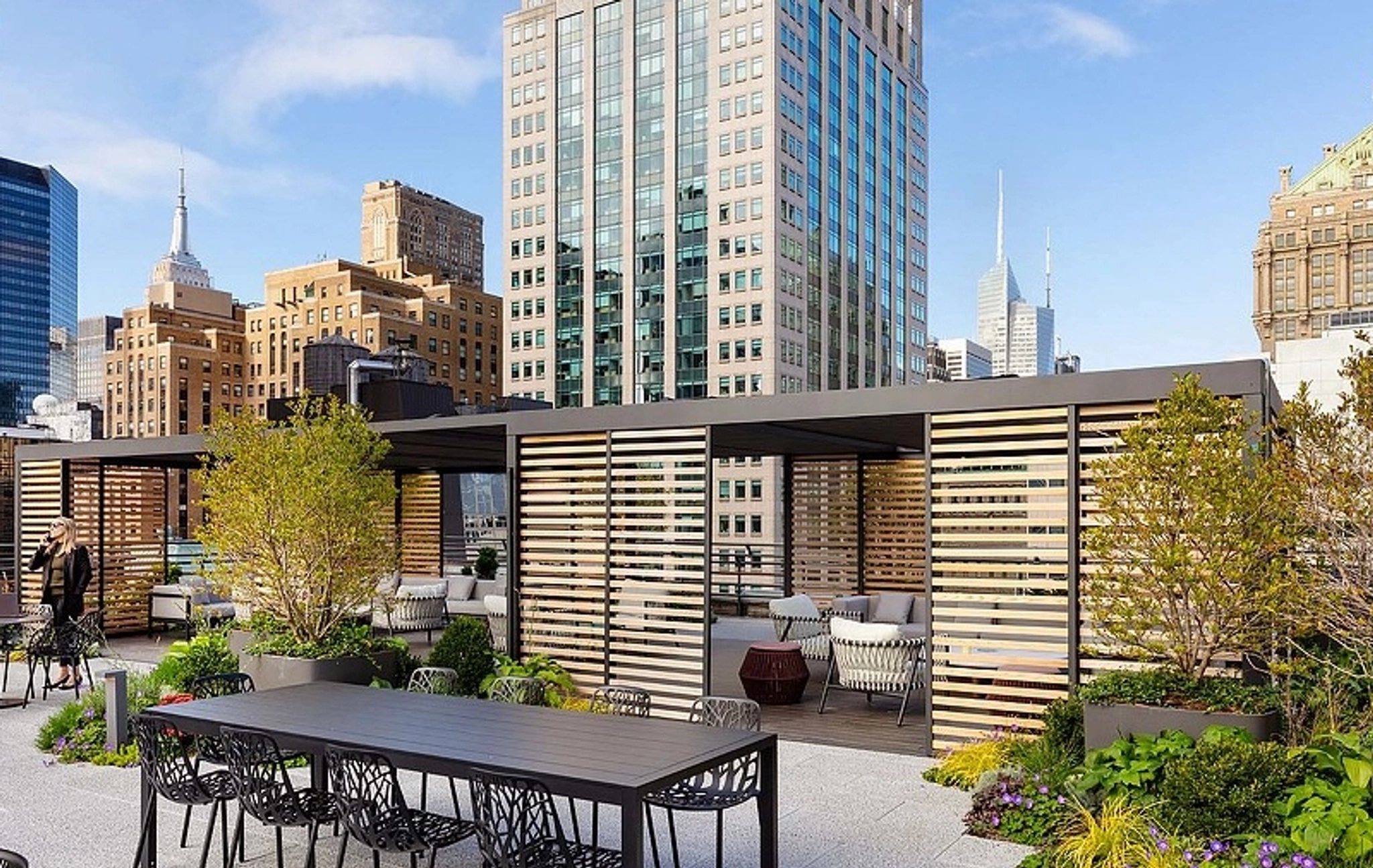 Rooftop terrace with seating and plants, set against a backdrop of tall buildings. This vibrant coworking area features a long table, chairs, and shaded seating areas, perfect for turning your workspace into an inspiring environment with city skyscrapers in the distance.