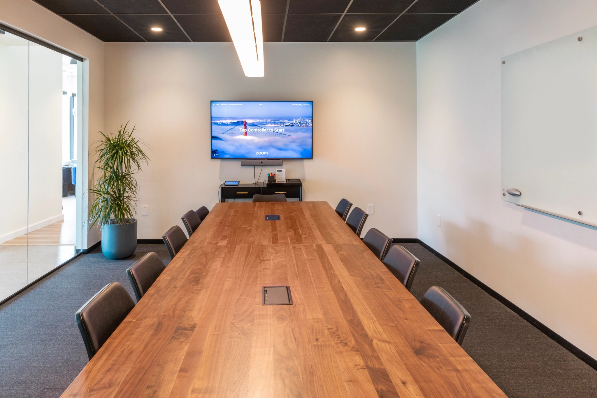 Een moderne coworking-ruimte in Chapel Hill beschikt over een vergaderruimte met een lange houten tafel, verschillende stoelen, een potplant, een aan de muur gemonteerd televisiescherm en een whiteboard.