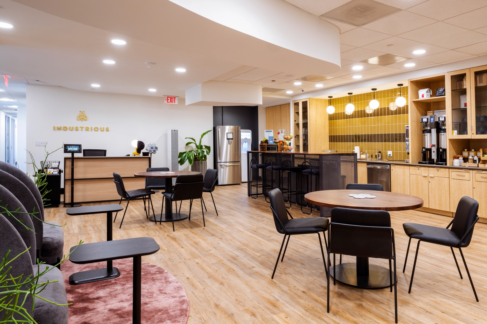 Moderne kantoorlounge in Boston met houten vloeren, ronde tafels, stoelen en een kitchenette met koffiezetapparaat en koelkast. Sfeerverlichting verbetert de sfeer op de werkvloer. ‘Industrious’ staat trots op de muur.