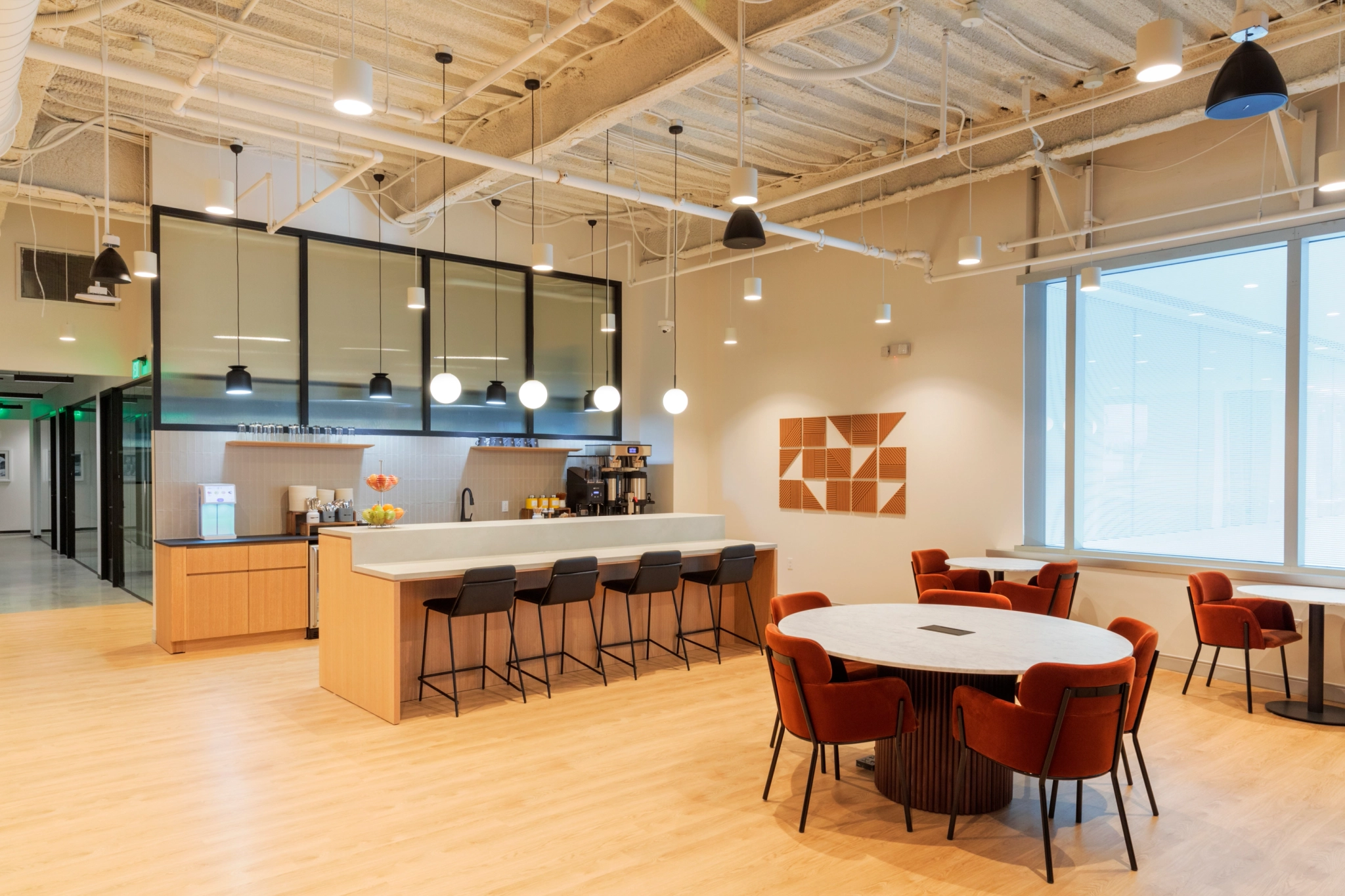 Eine moderne Coworking-Küche in Jersey City verfügt über eine elegante Theke mit Barhockern, runde Tische mit Stühlen und stilvolle Hängelampen. Beige Wände und heller Holzboden runden die einladende Büroatmosphäre ab.