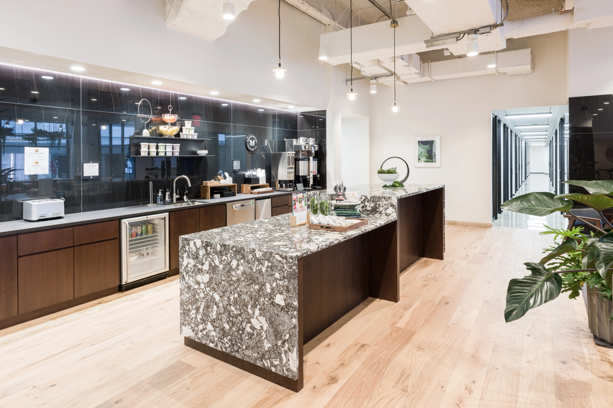 Una moderna cocina de oficina en Filadelfia cuenta con una isla de mármol, gabinetes de madera, cafeteras y luces colgantes. Plantas y una pieza decorativa decoran la isla. Un pasillo a la derecha conduce a la sala de reuniones.