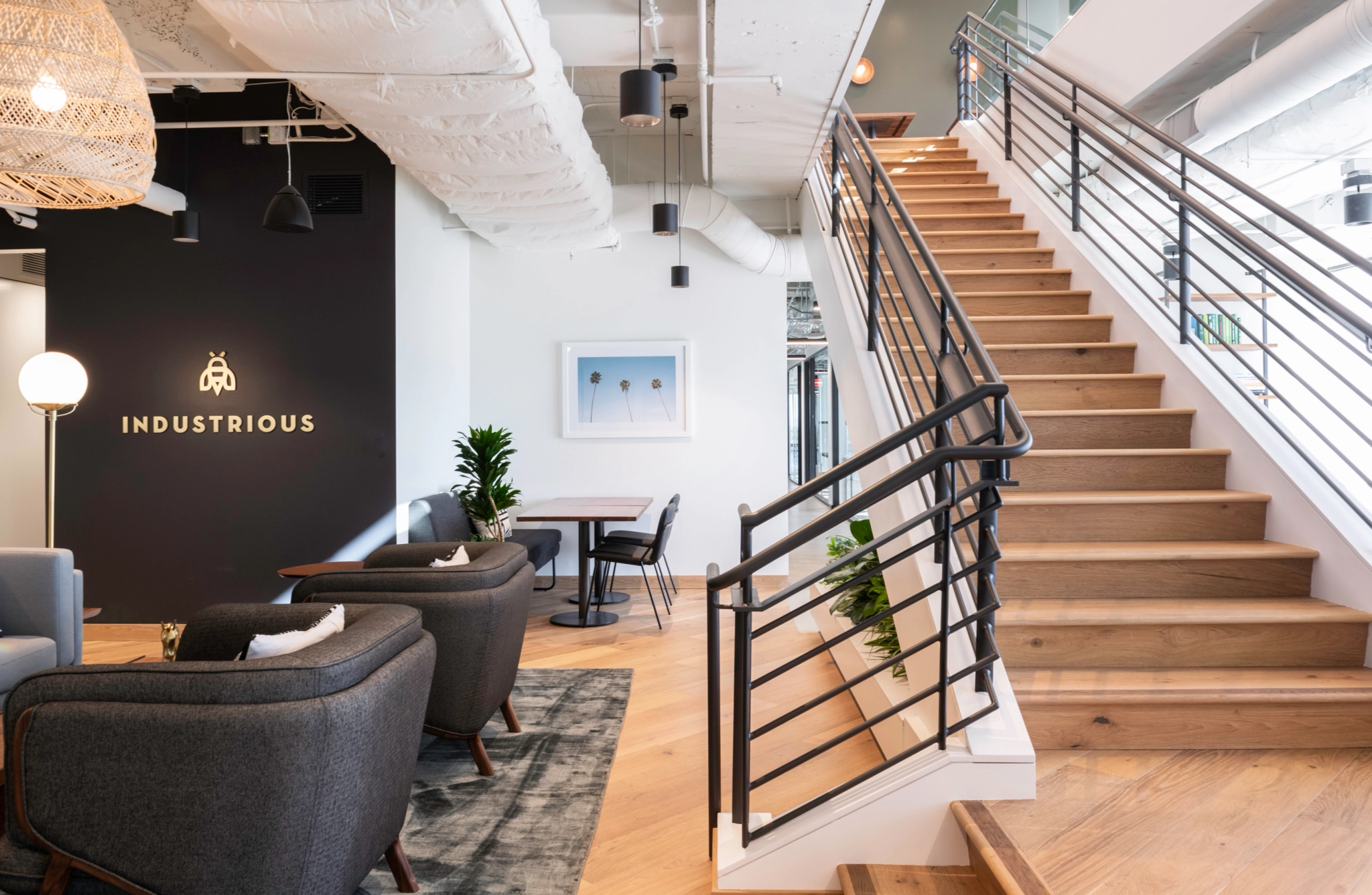 Moderno espacio de oficina de coworking en Pasadena con una escalera, sillas negras, una mesa de madera, plantas y una pared que muestra con orgullo la palabra "INDUSTRIOUS".