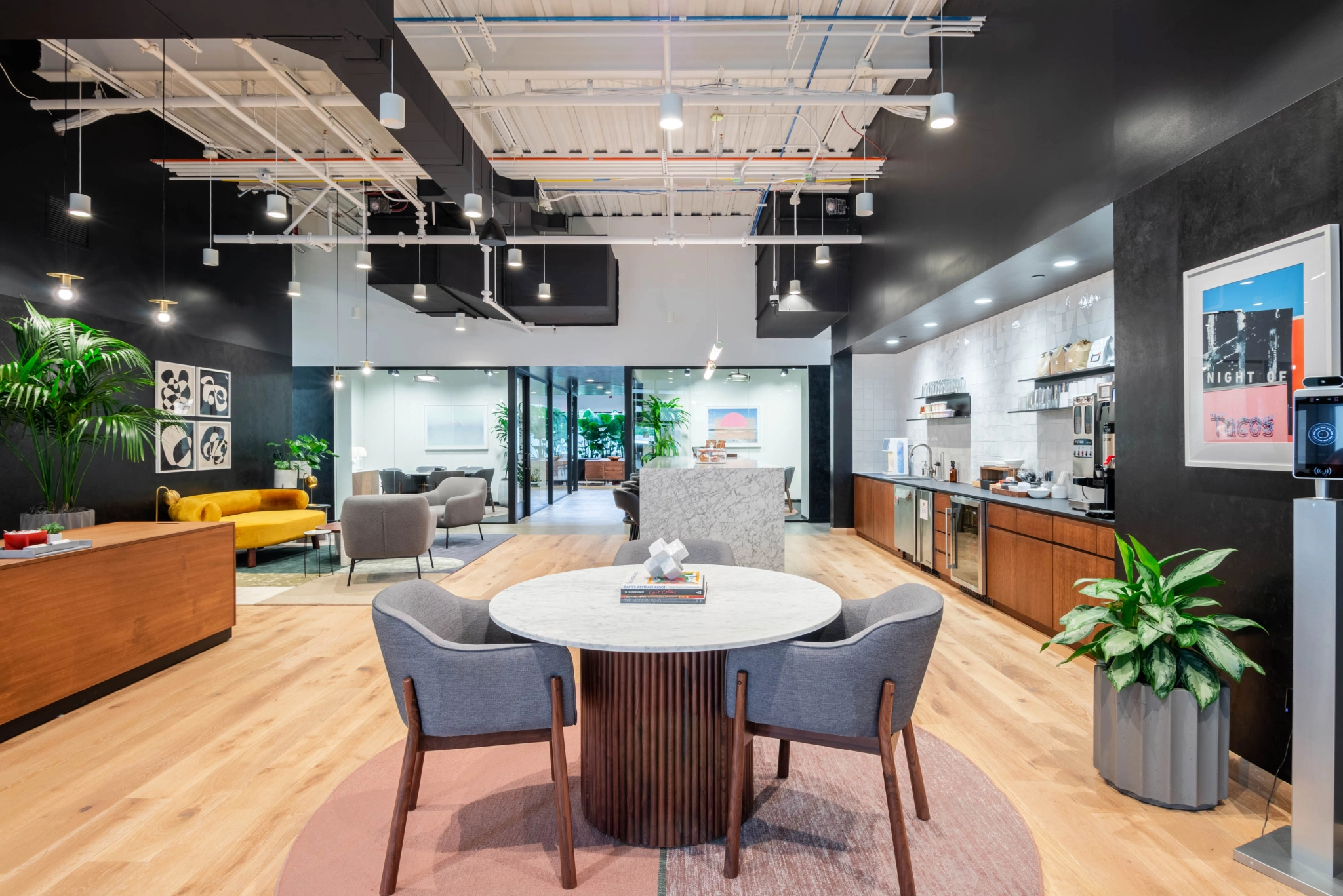 Salón de oficina moderno en Short Hills con mesa redonda y sillas, cómodos sofás, vibrantes obras de arte en las paredes y plantas exuberantes. A la derecha hay un área de cocina, mientras que al fondo una sala de reuniones con paredes de vidrio complementa el dinámico ambiente de coworking.