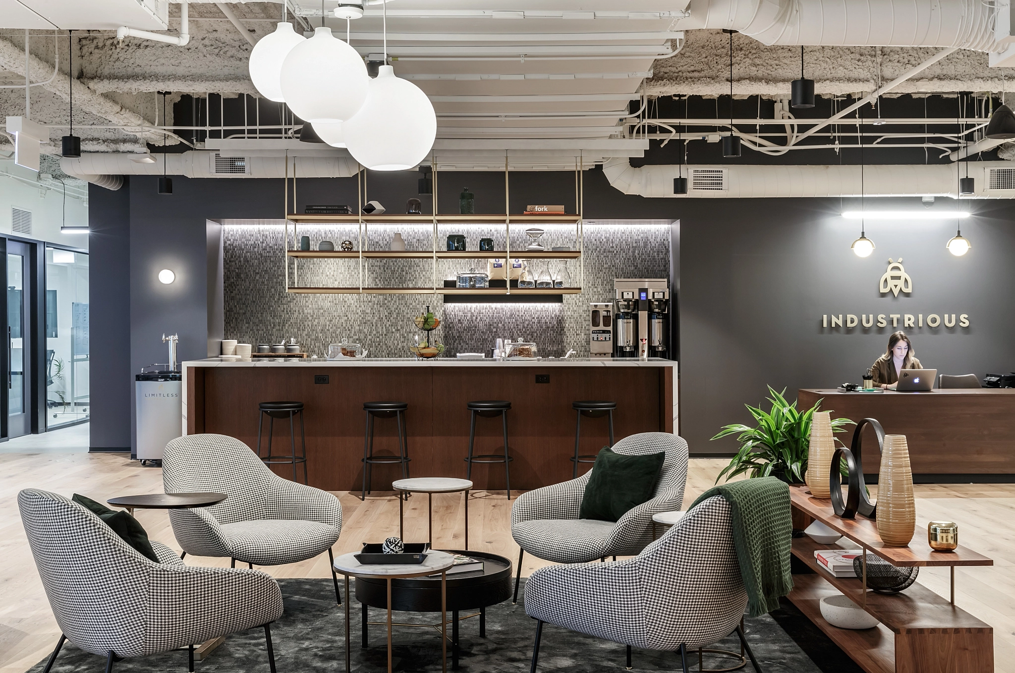 Moderne Bürolounge mit weichen Stühlen, einer Kaffeebar und einer Frau, die an der Rezeption arbeitet. Das „Industrious“-Logo schmückt die Wand, während Hängelampen und Dekor das zeitgenössische Design unterstreichen. Dieser Raum ist ideal für Coworking in Chicago und verfügt außerdem über einen angrenzenden Besprechungsraum für alle Ihre Bedürfnisse.