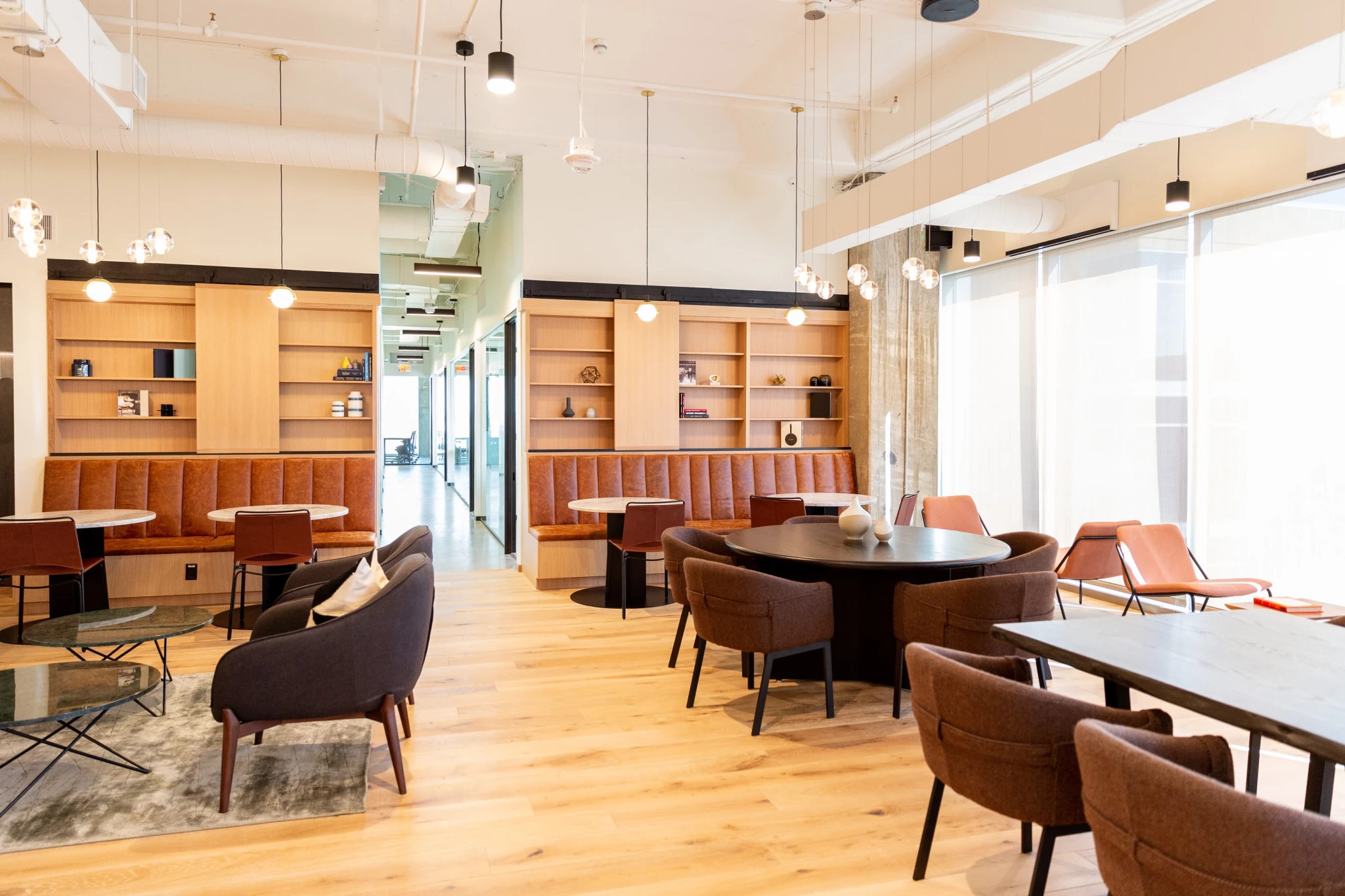 Un moderno salón de coworking está amueblado con mesas redondas, sillas marrones y zonas para sentarse. Los estantes se alinean en las paredes, mientras que las lámparas colgantes iluminan el espacio, que está inundado de luz natural que entra a través de los grandes ventanales, un escenario perfecto para reuniones colaborativas.