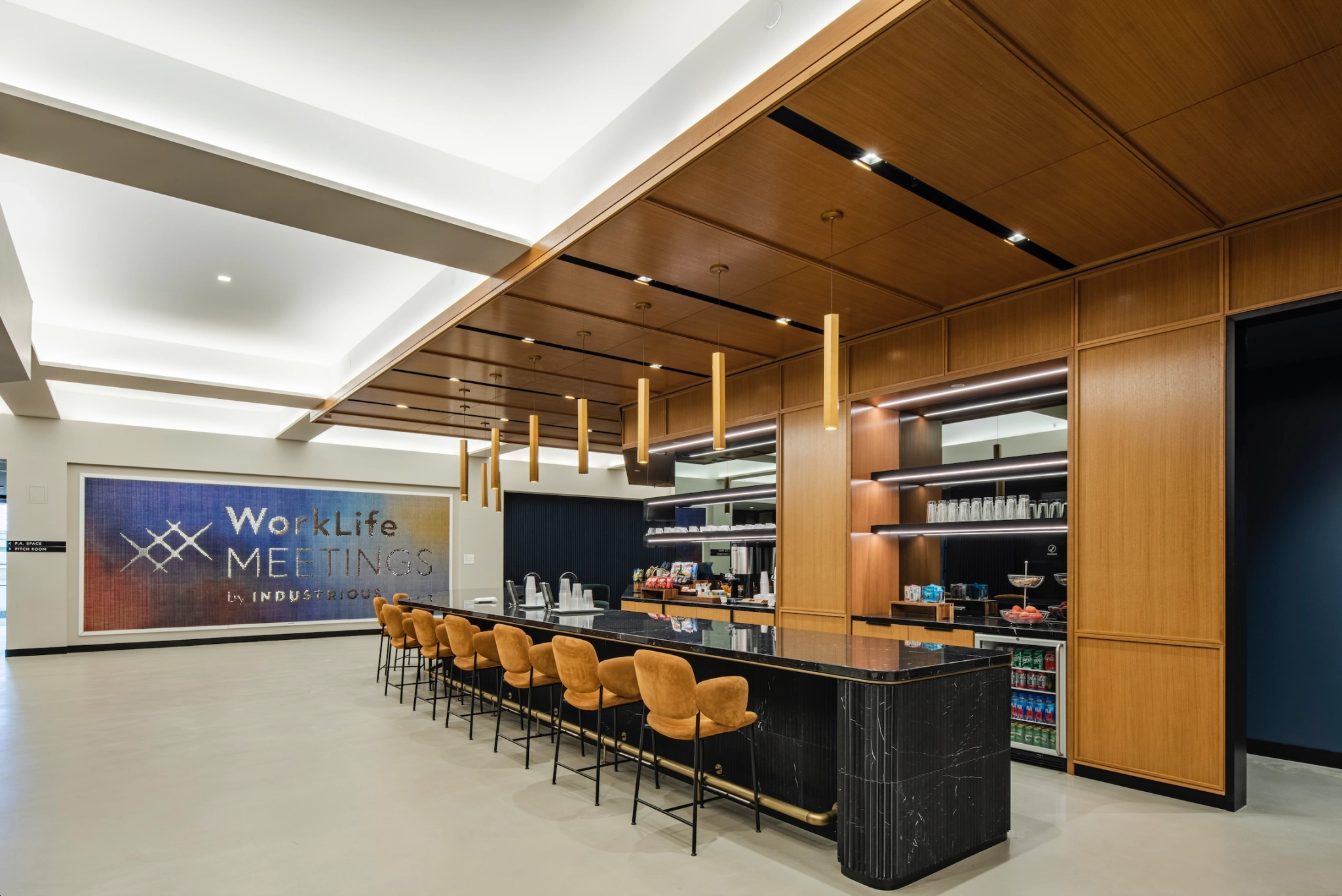 Modern New York office kitchen and lounge area with a long black counter, multiple chairs, wooden cabinetry, overhead lighting, and a sign displaying "WorkLife Meetings by Industrious.