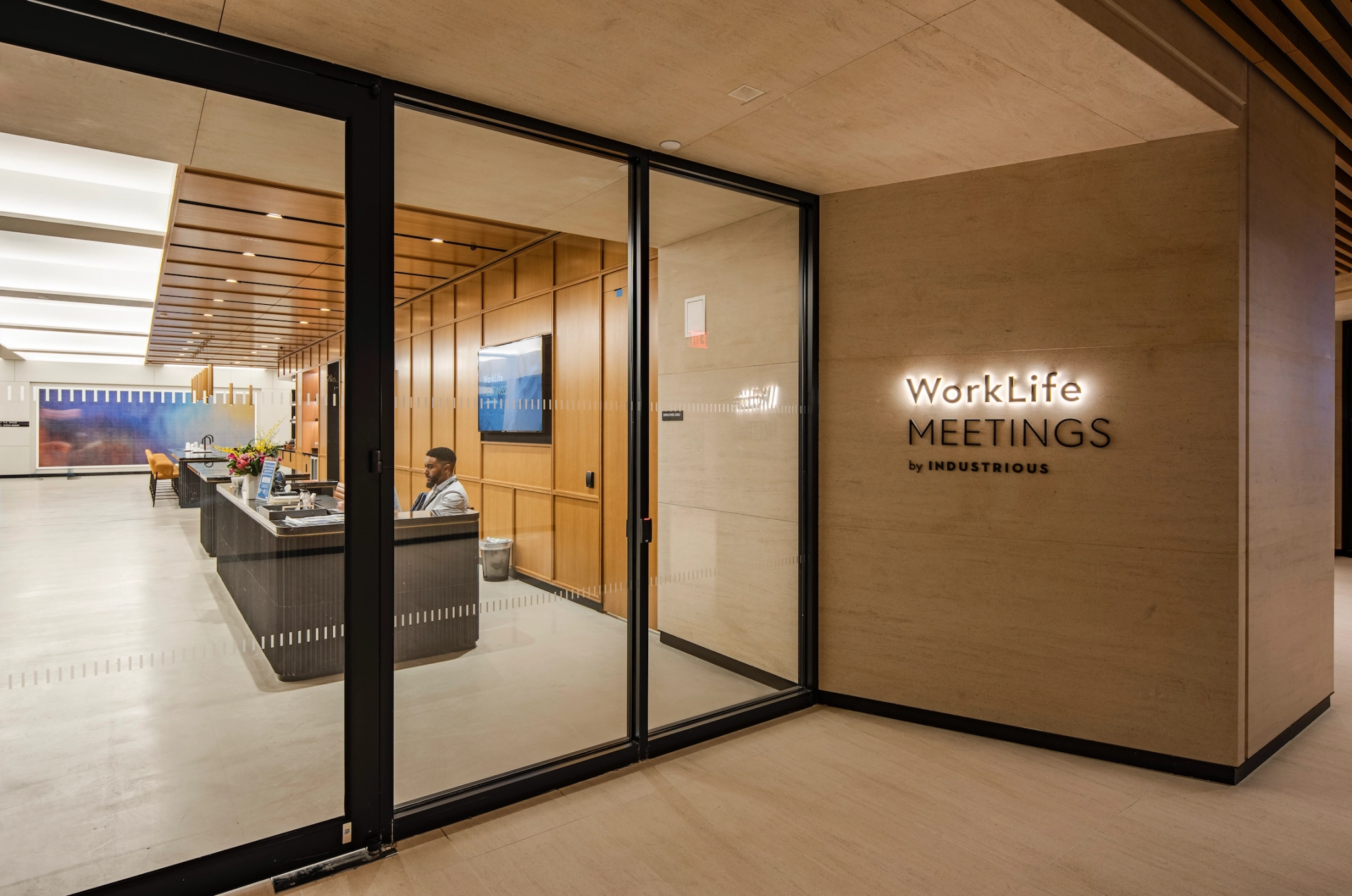 A modern office lobby with a reception desk, glass doors, and a sign reading "WorkLife Meetings by Industrious" on the wall. An individual is seated at the desk in this New York workspace.