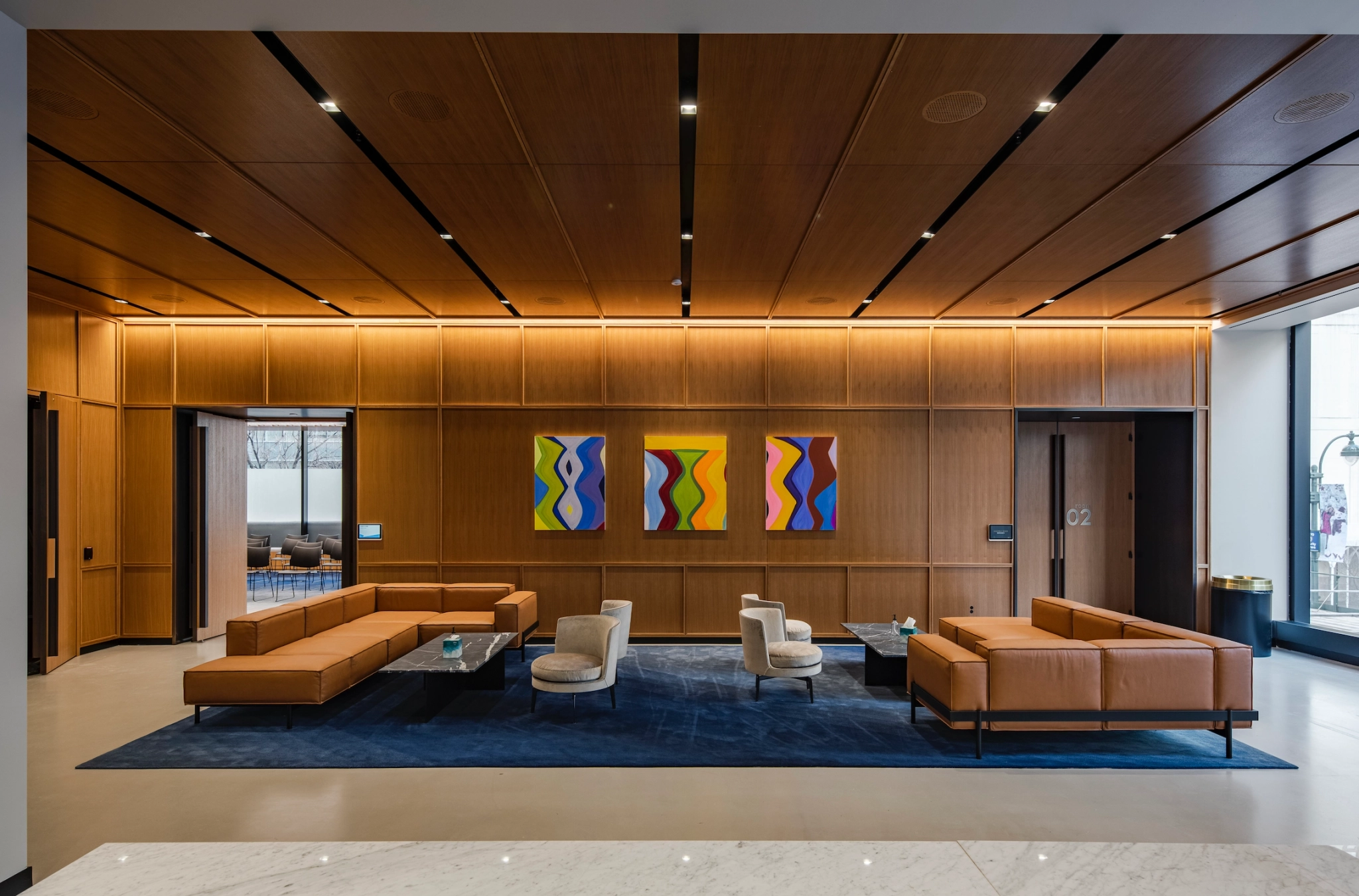 A modern coworking lobby with wooden panel walls, three colorful abstract paintings, brown leather seating, and a dark blue rug. Large windows on the right let in natural light.