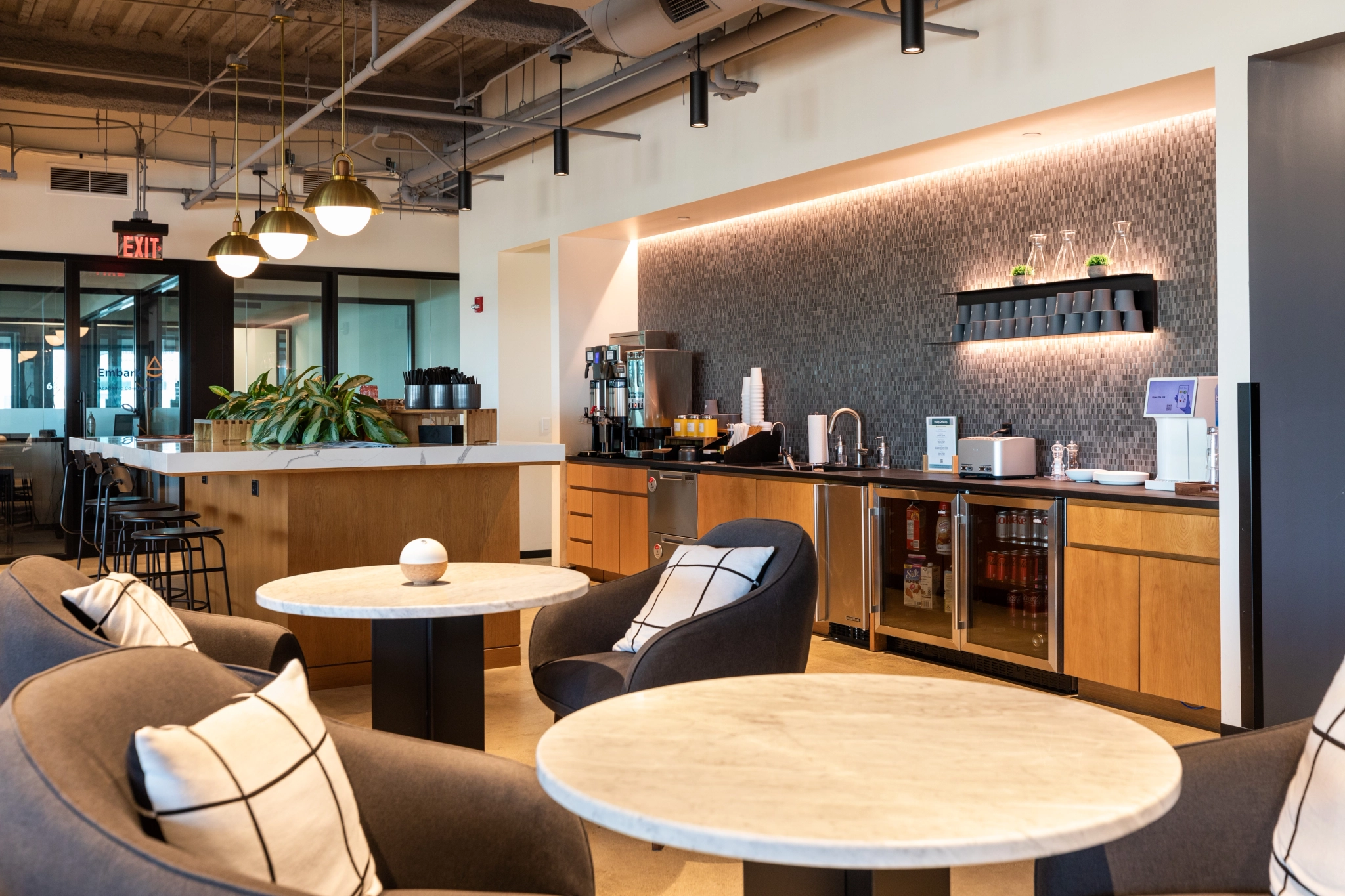 Diese Coworking-Lounge in Evanston verfügt über moderne runde Tische, gepolsterte Stühle, eine Kaffeestation und Sitzgelegenheiten in der Bar. Der Arbeitsbereich wird durch Pflanzen und eine strukturierte Wand aufgewertet, während Deckenleuchten den Bereich warm erhellen.