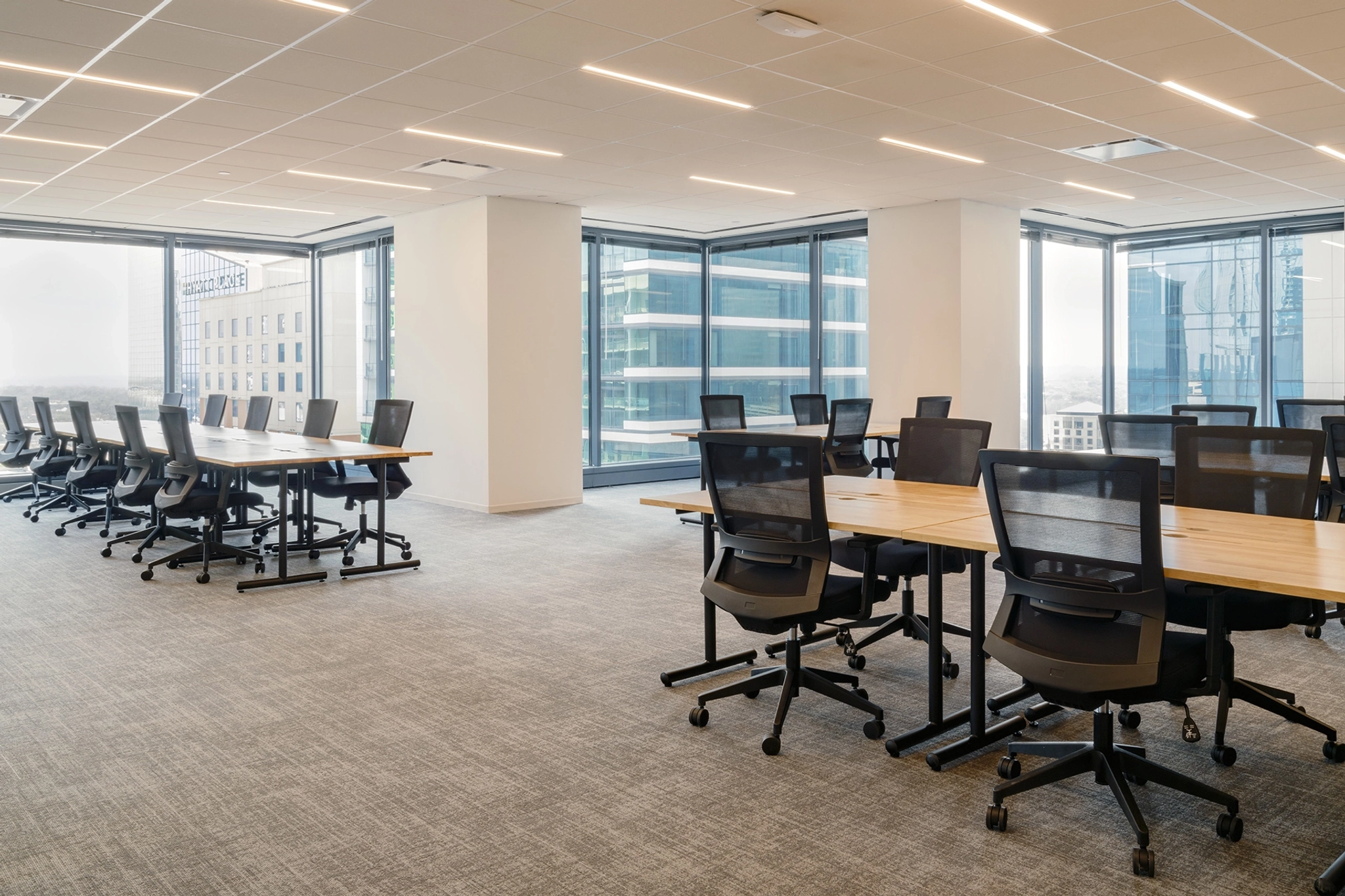 Espace de bureau moderne avec des rangées de tables et de chaises vides, un éclairage vif et de grandes fenêtres donnant sur la ville, conçu comme un espace de travail polyvalent.