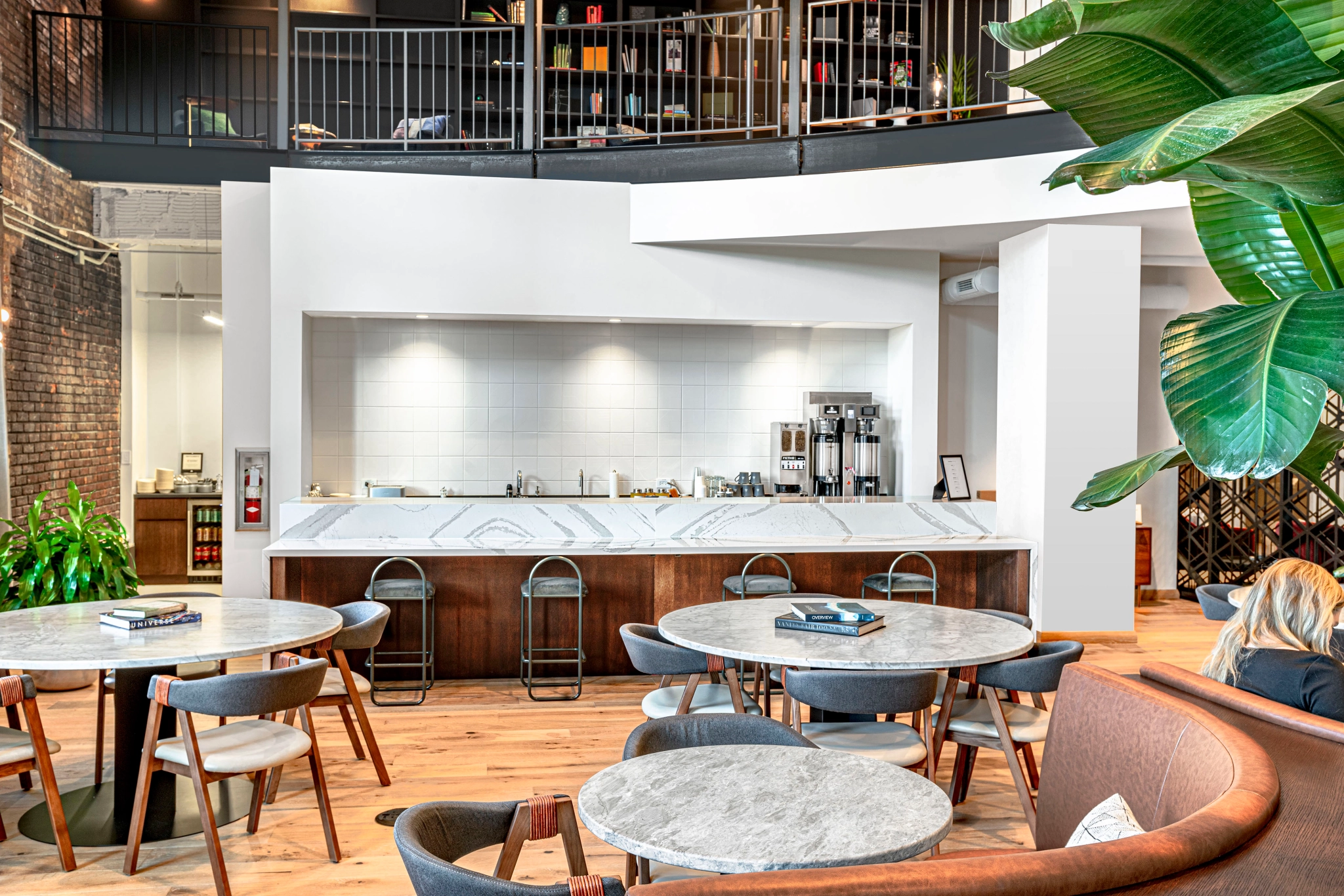 El interior de una cafetería moderna en Chicago con mesas y sillas redondas de mármol junto a un elegante mostrador blanco. Ideal como un acogedor espacio de coworking, decorado con plantas verdes y con un encantador balcón arriba.