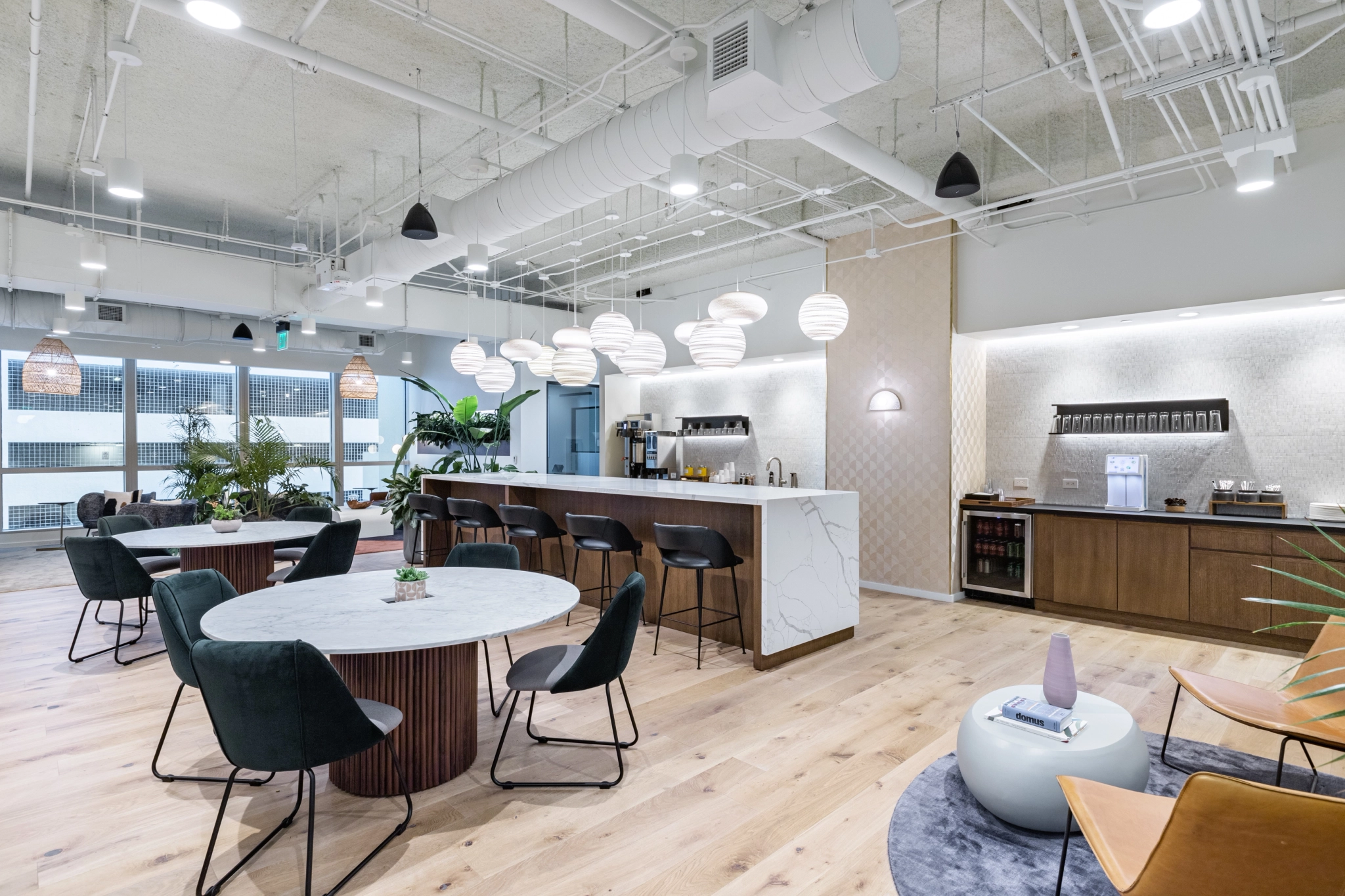 Un salón de coworking moderno con cocina americana, mesas redondas, sillas negras, encimeras blancas y lámparas colgantes. Los grandes ventanales ofrecen vistas vibrantes de la ciudad de Miami.