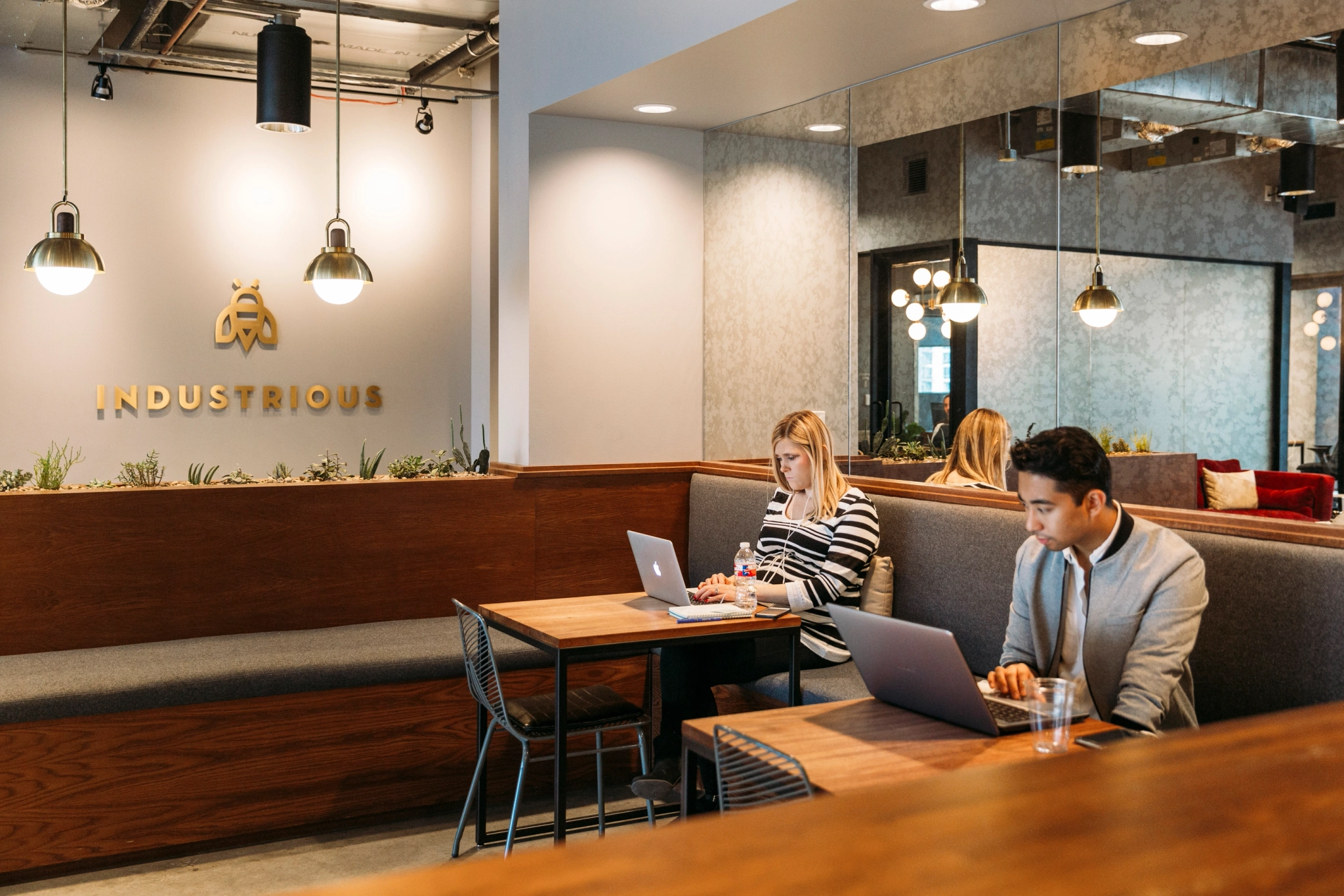 Twee mensen werken op laptops in een moderne coworking-ruimte in Austin. De werkruimte is voorzien van stoelen met kussens, hanglampen en een bord met de tekst 'Industrious' aan de muur.