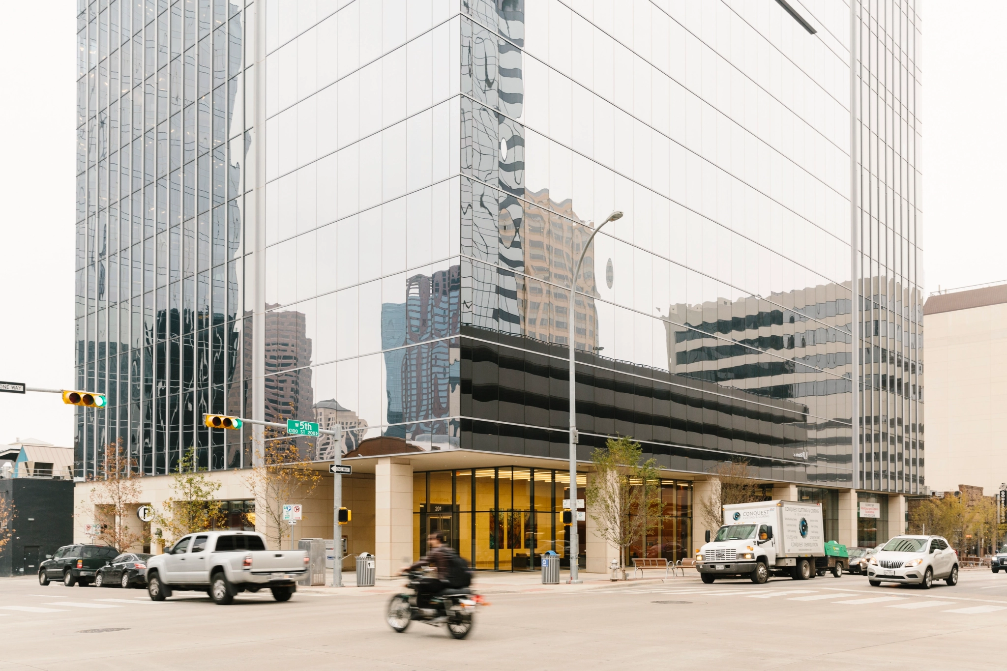 Una concurrida intersección en Austin con vehículos que incluyen una motocicleta, automóviles y un camión frente a un edificio de oficinas hecho de vidrio reflectante. También se pueden ver semáforos y señales de tráfico.