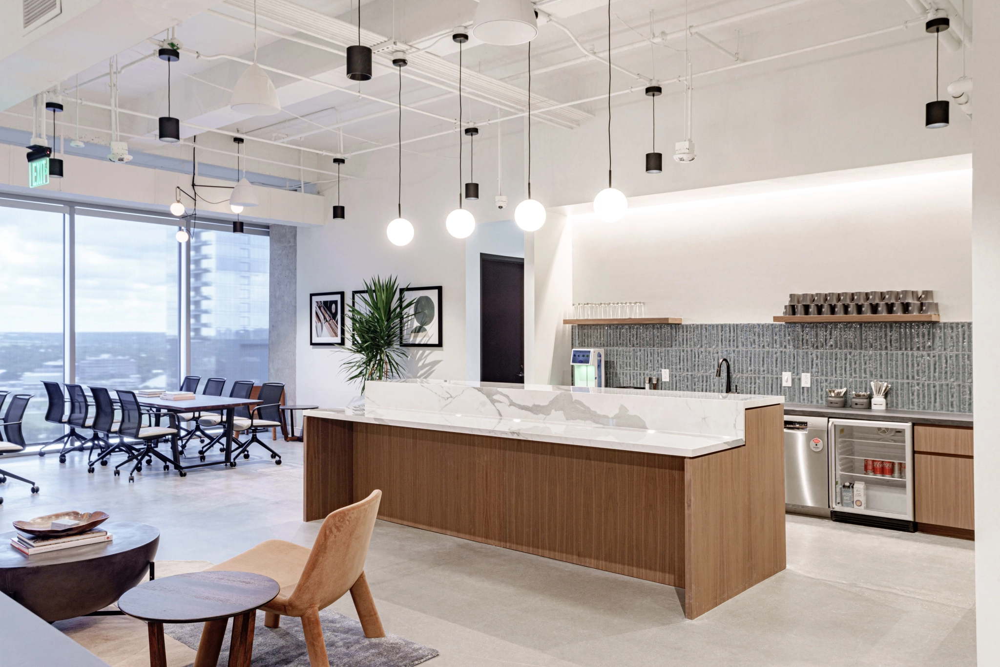 Modern coworking office interior with a spacious kitchen area featuring a marble countertop, hanging lights, and a meeting room with a table and chairs by the window.