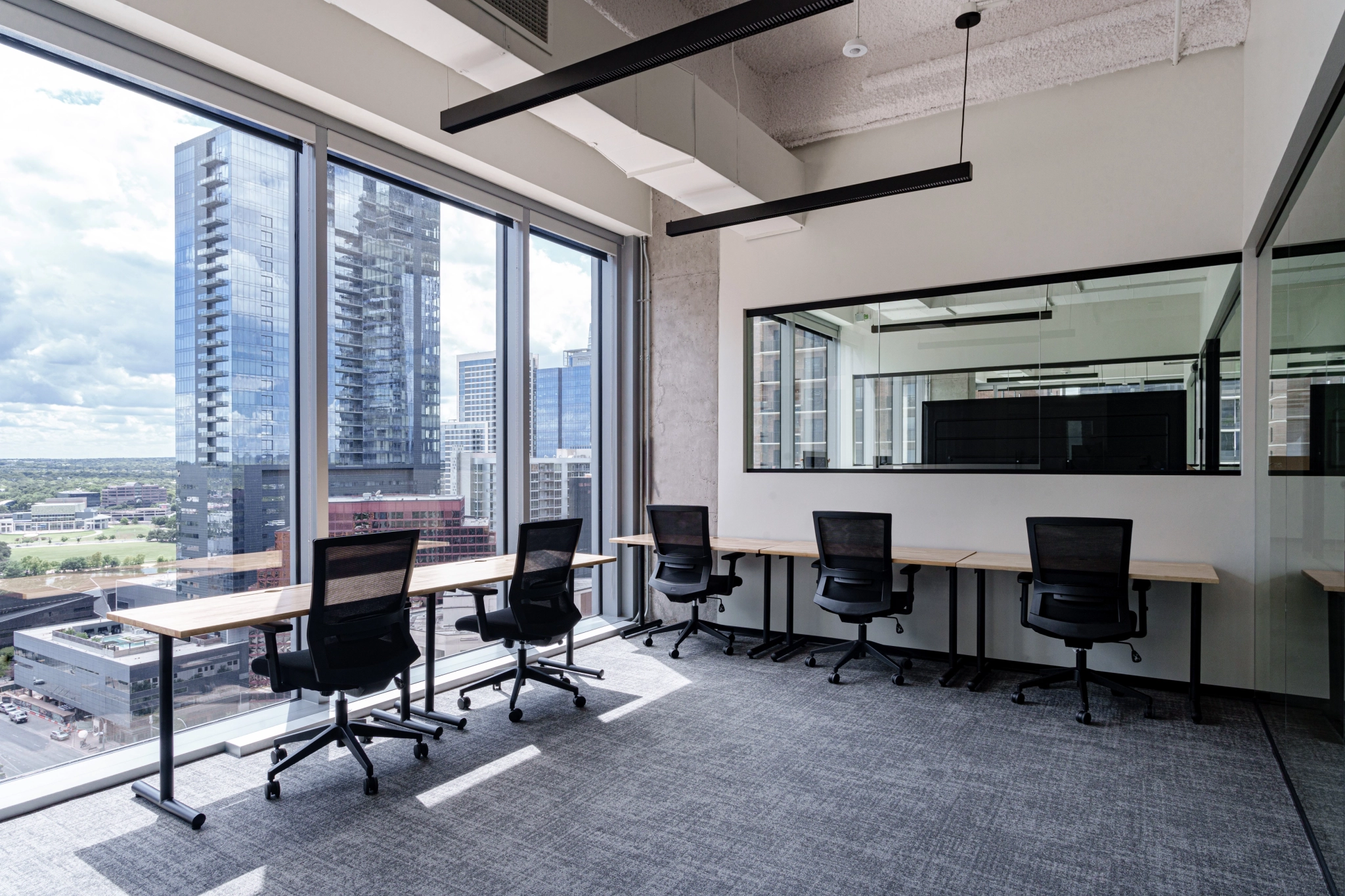 Modern workspace with large windows, four desks, and chairs, overlooking city skyscrapers.