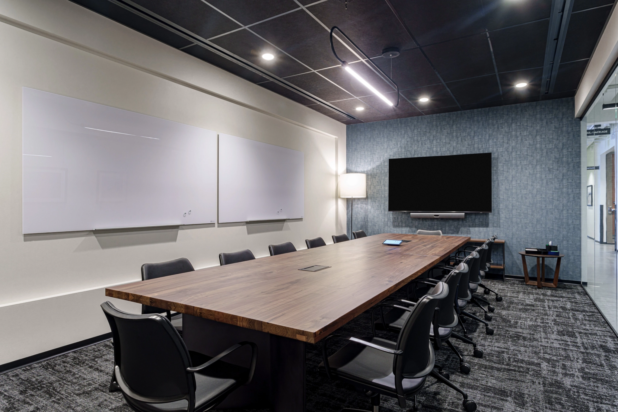 Une salle de conférence moderne à Austin dispose d'une longue table en bois entourée de chaises de bureau. Elle comprend une télévision à écran plat, des tableaux blancs et un lampadaire, le tout sur un plafond sombre et un sol recouvert de moquette, créant ainsi un espace de travail efficace.