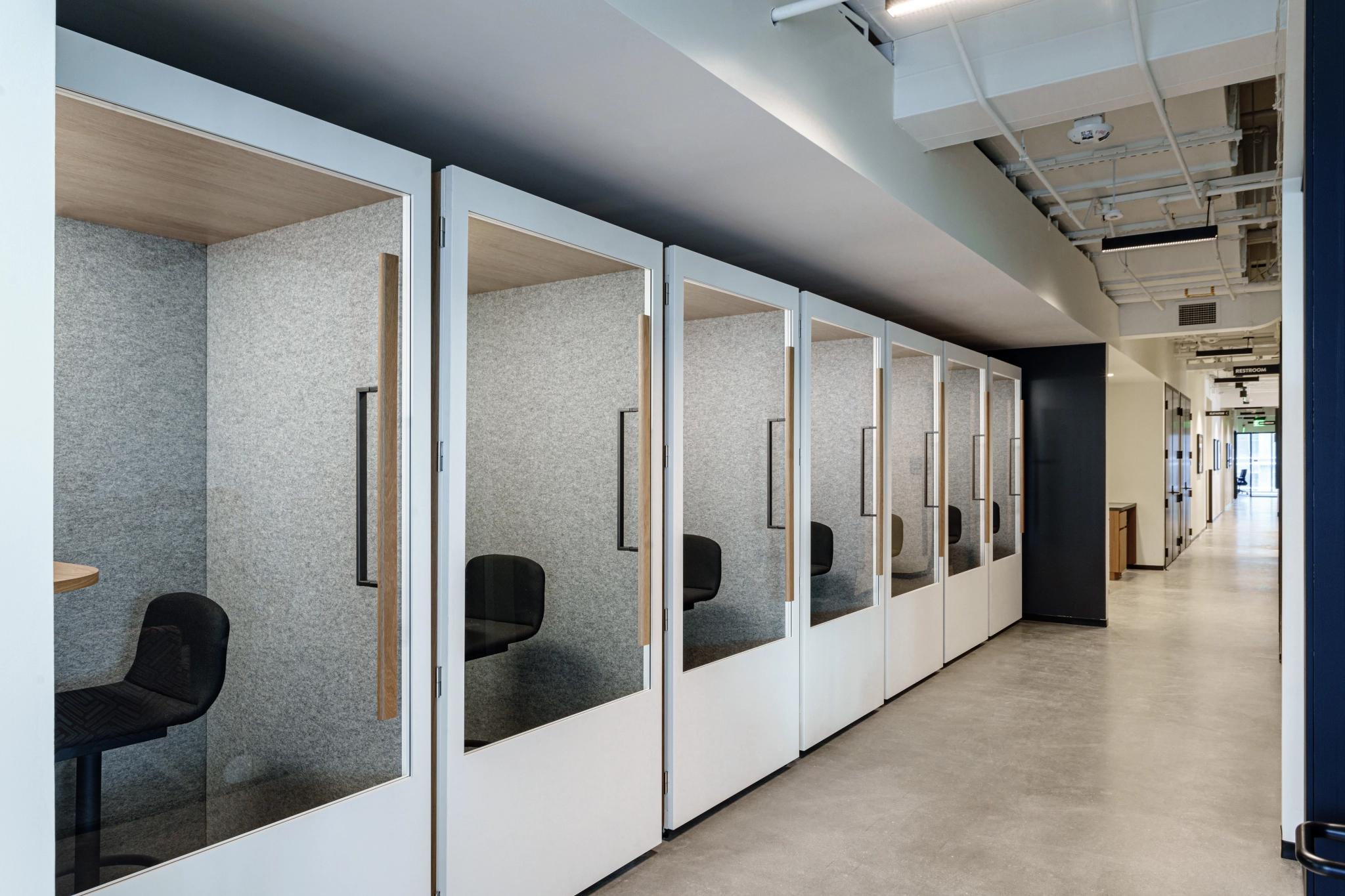 A row of modern office phone booths with glass doors and chairs is set along a hallway in a coworking space, featuring a concrete floor and exposed ceiling.