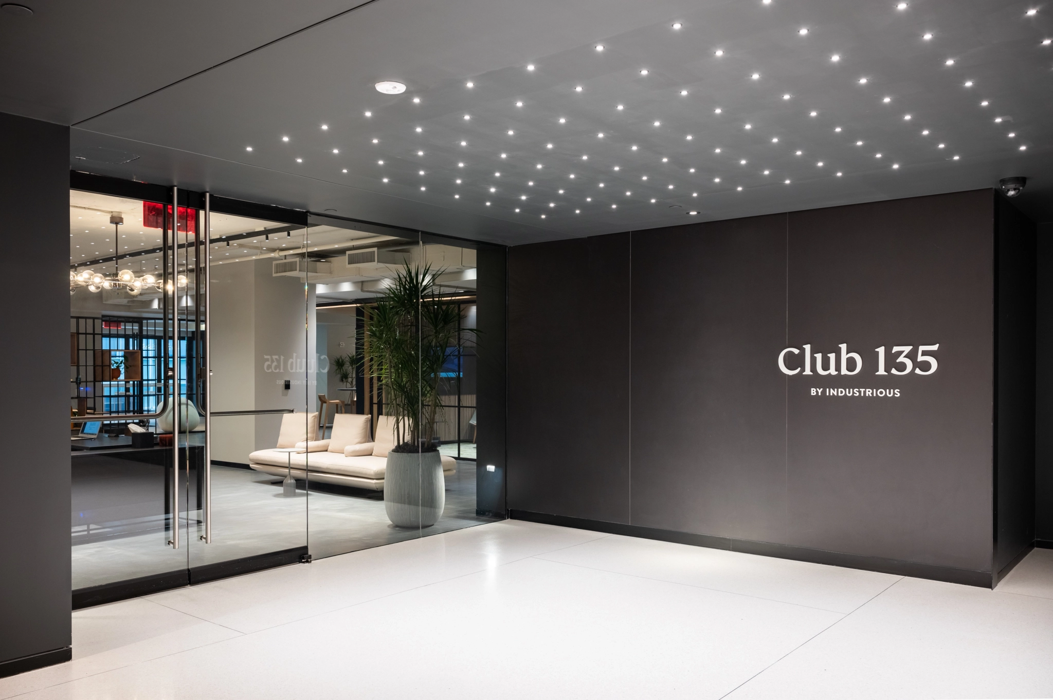 Modern office lounge area with the sign "Club 135 by Industrious" on a dark wall, illuminated by ceiling lights, and furnished with sofas and a plant near glass doors, seamlessly blending into the adjacent coworking workspace.