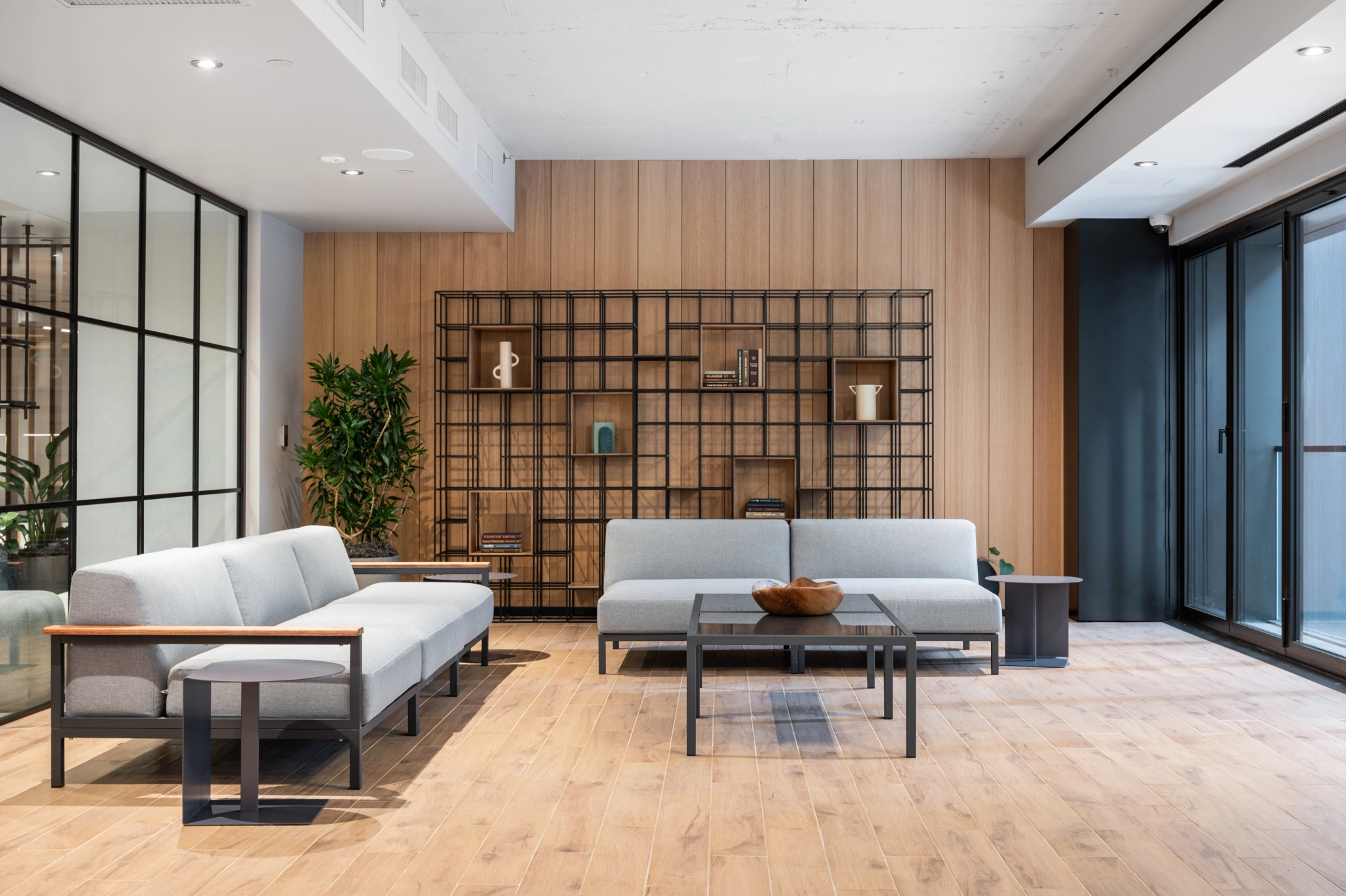 A modern, minimalist living room in New York with light gray sofas, a wooden coffee table, a metal-framed shelf with decor items, wooden flooring, and large windows providing natural light. The versatile space easily doubles as an elegant workspace.