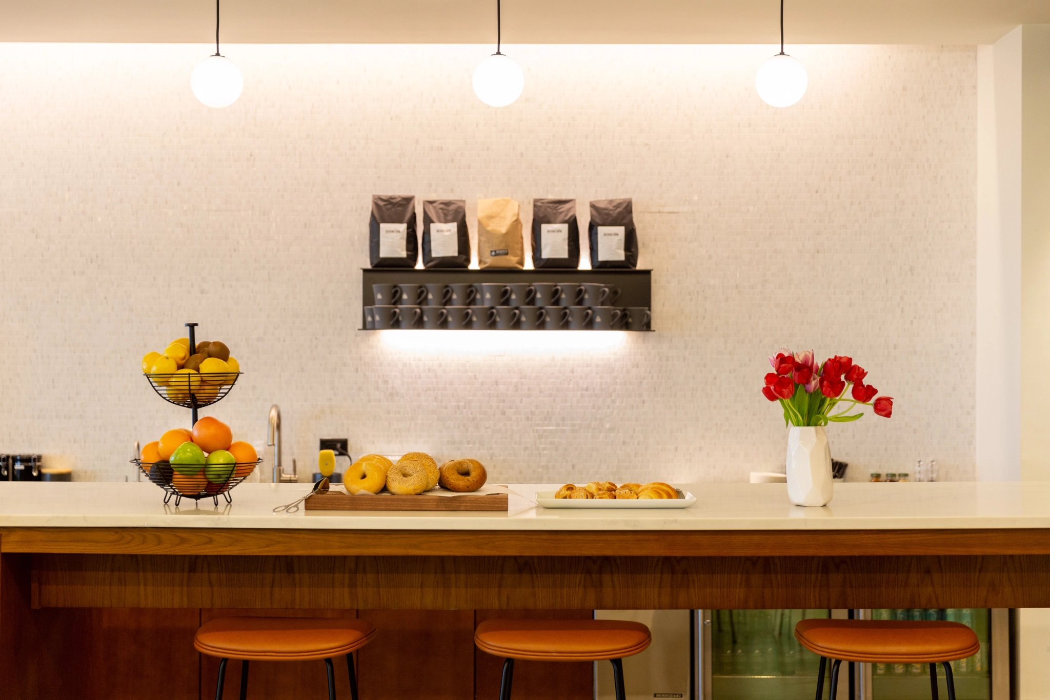 Una canasta de frutas de tres niveles, una bandeja de bagels y pasteles y un jarrón blanco con tulipanes rojos se encuentran en el mostrador de la cocina en la sala de conferencias. Al fondo, estantes con bolsas de café y tres luces colgantes, crean una atmósfera acogedora para sesiones informales de coworking.