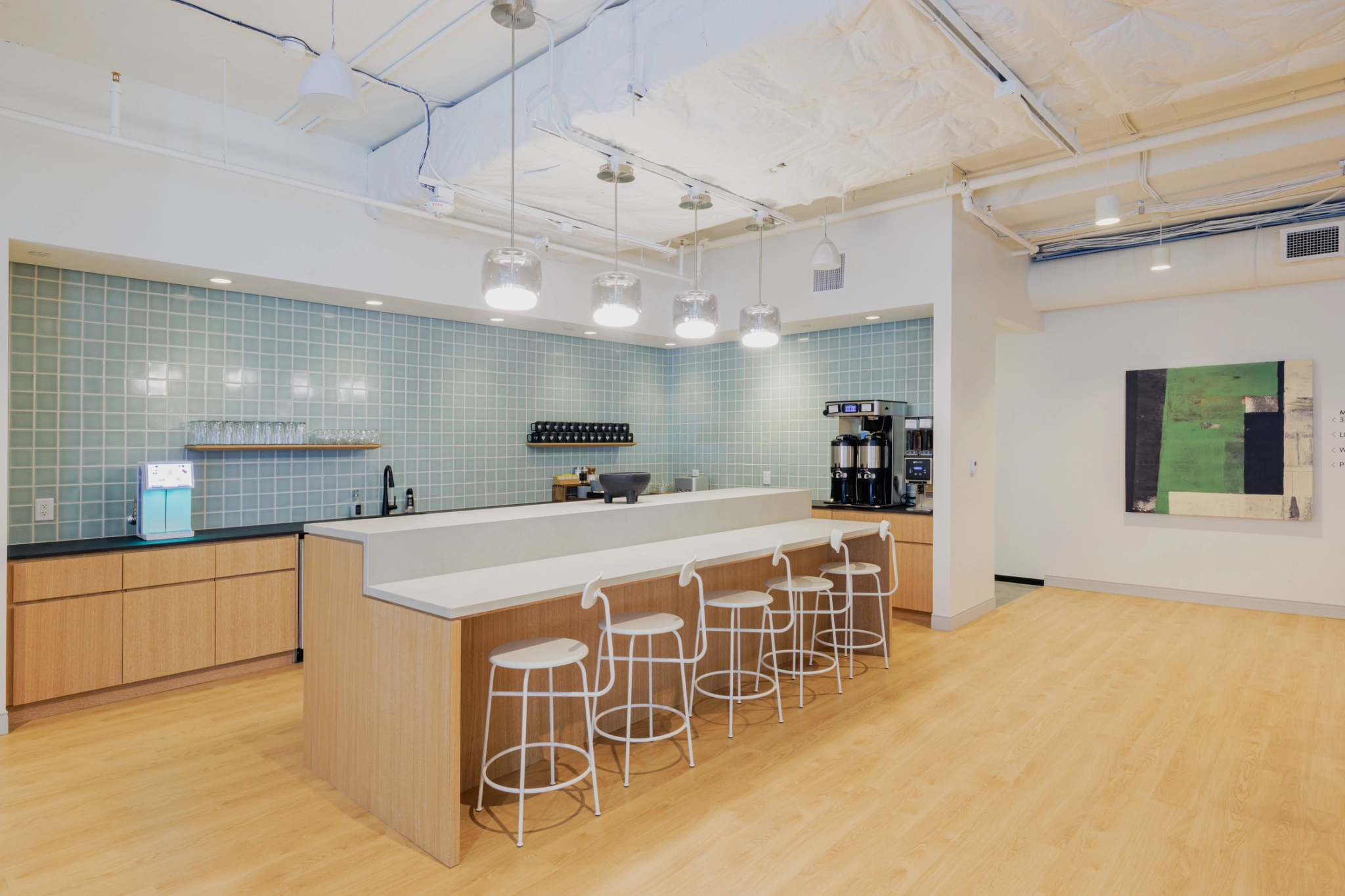 Una cocina moderna con una barra larga, taburetes, máquinas de café y una pared trasera con azulejos azules es el lugar de trabajo perfecto en la oficina. Las luces del techo cuelgan sobre el mostrador y una obra de arte abstracta cuelga de la pared adyacente para inspirarse.