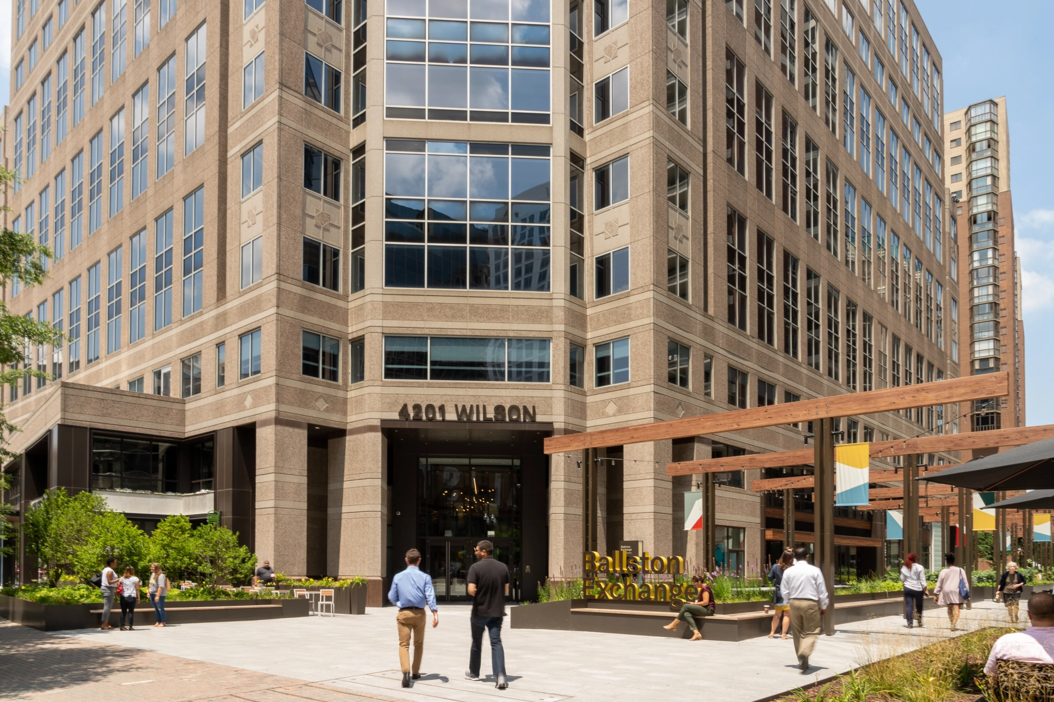 Un edificio de oficinas de varios pisos con letreros "4201 Wilson" y espacios de coworking. La gente pasa por la entrada y la zona de asientos al aire libre marcada como "Ballston Exchange". Varias tiendas y espacios verdes rodean la concurrida zona de trabajo.