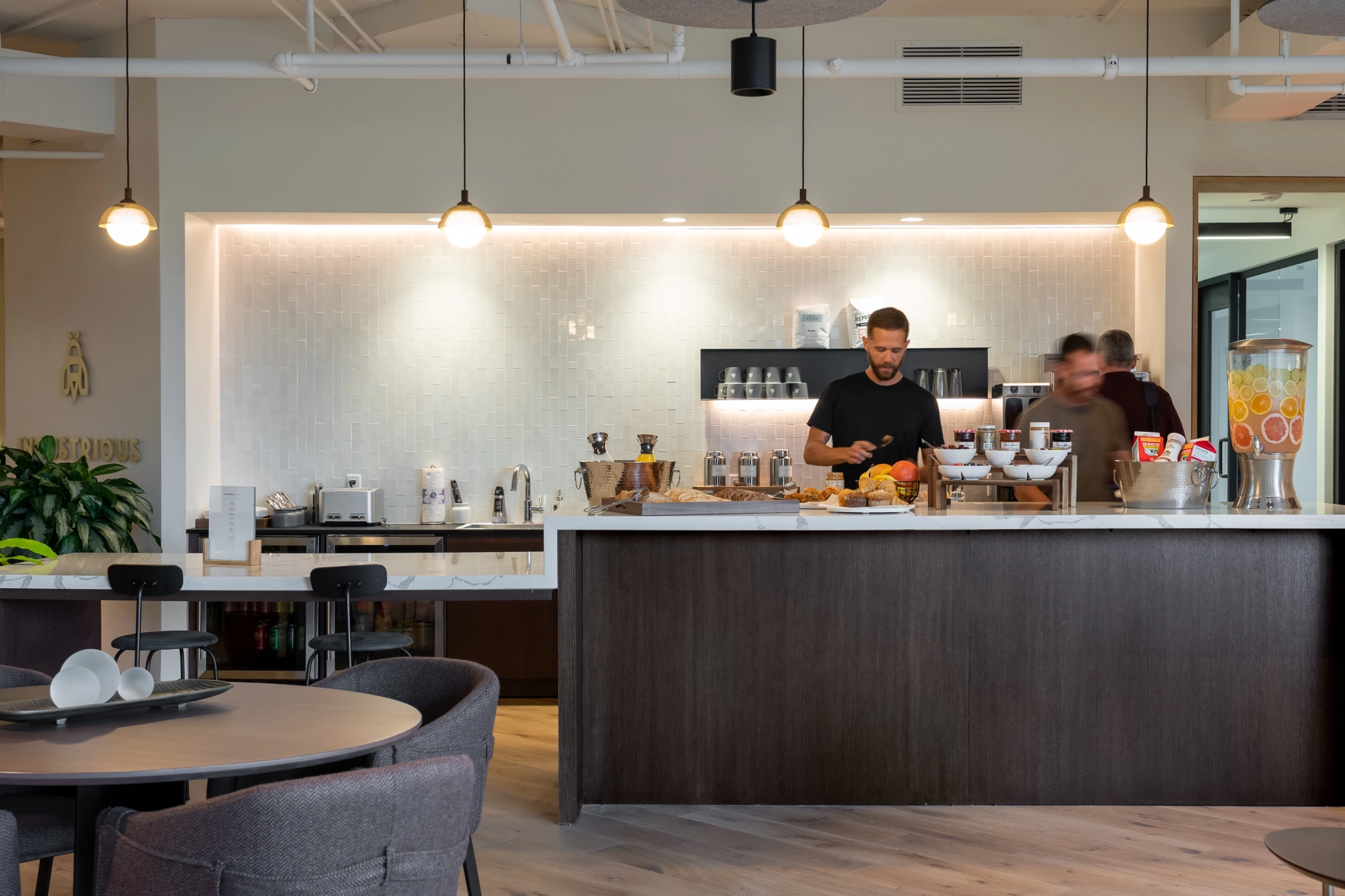 In een moderne keuken met een witte betegelde achterwand bereiden twee mensen eten achter een donkerhouten aanrecht. De een concentreert zich op taken, terwijl de ander zich in een waas beweegt, alsof hij in een coworkingkantoor zit. Er is een sapdispenser aan de rechterkant, wat bijdraagt aan de efficiënte sfeer.