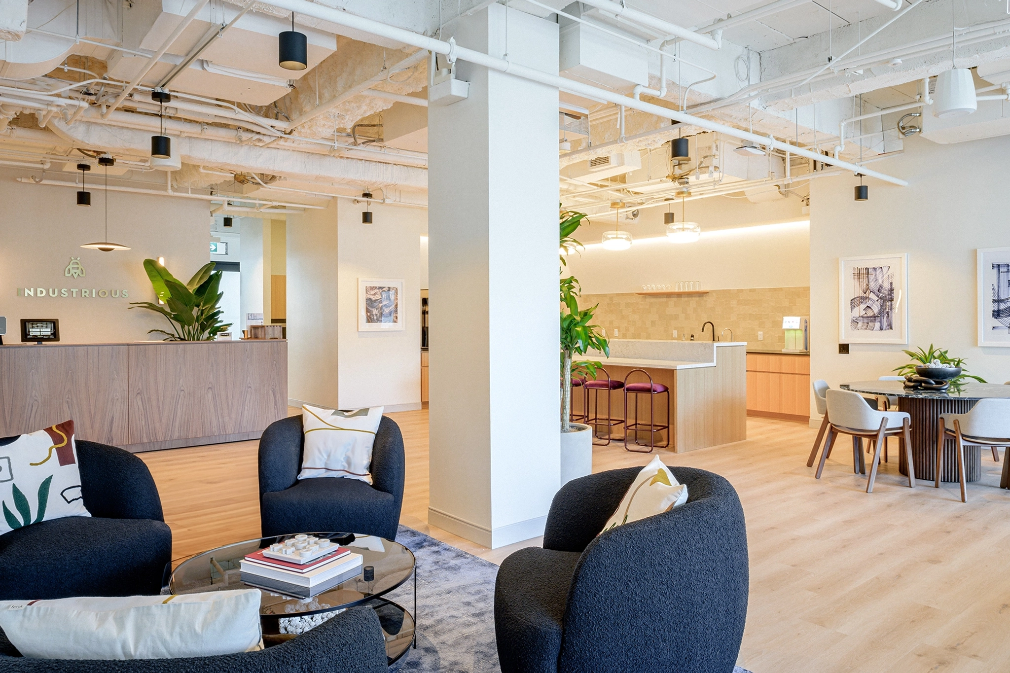 El moderno salón de oficina, perfecto para coworking, ofrece cómodos asientos y una mesa pequeña, con una cocina al fondo. La habitación cuenta con suelo de madera clara, paredes blancas y techo con tuberías a la vista.