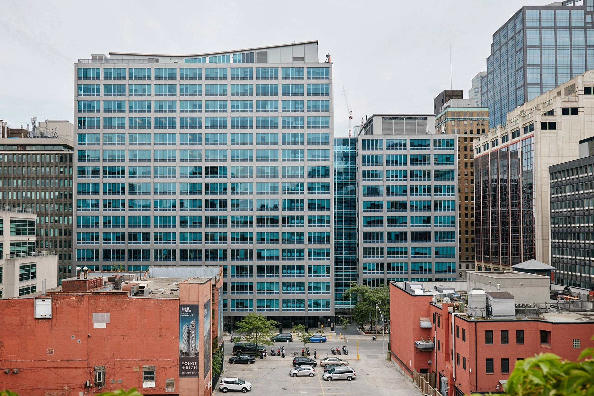 Un gran edificio de oficinas moderno con ventanas de cristal azul, idealmente situado en Toronto, está flanqueado por otros edificios. Varios coches están aparcados en un aparcamiento delante de la puerta, lo que indica la bulliciosa actividad en las numerosas salas de reuniones.