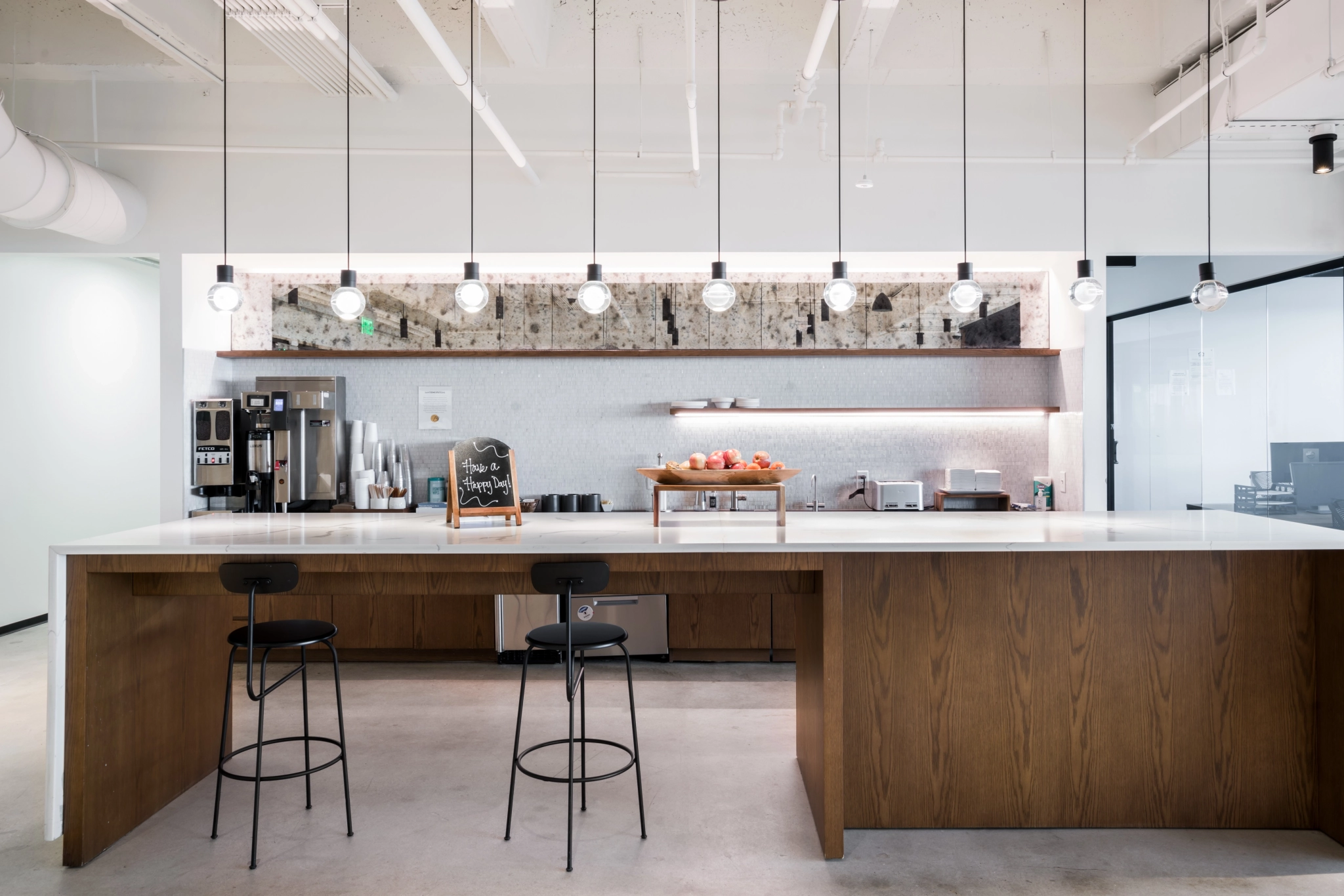 Una cocina moderna en Atlanta con una larga isla de madera, dos taburetes, luces colgantes, una estación de café y una pizarra en el mostrador. El telón de fondo es una pared de azulejos con estantes y electrodomésticos de cocina, que crea un espacio de trabajo acogedor para reuniones culinarias.