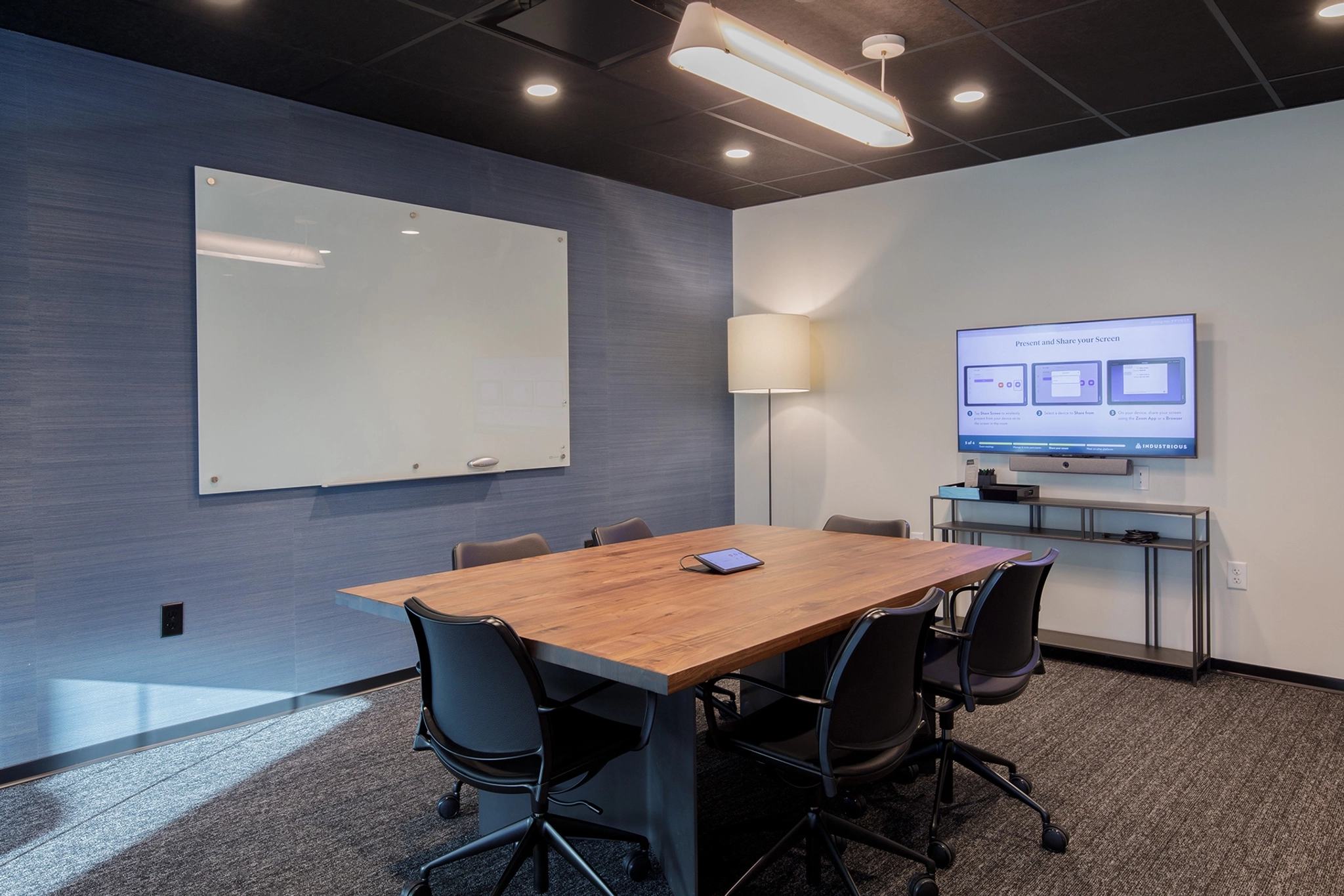 A modern conference room in Fort Lauderdale featuring a large wooden table, six chairs, a whiteboard, a mounted screen displaying a presentation, and a floor lamp provides an ideal workspace.