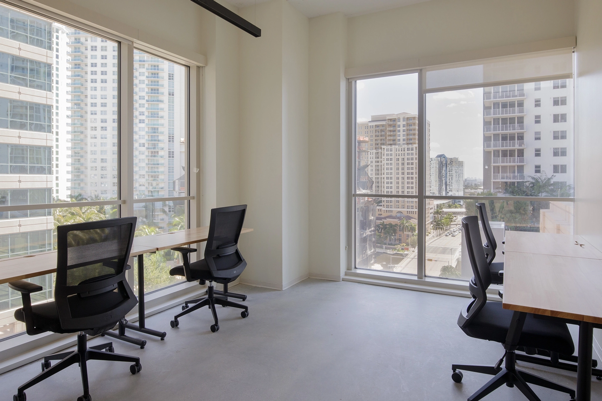 A modern workspace with several empty desks and ergonomic chairs, featuring large windows that offer a view of high-rise buildings and the Fort Lauderdale cityscape.