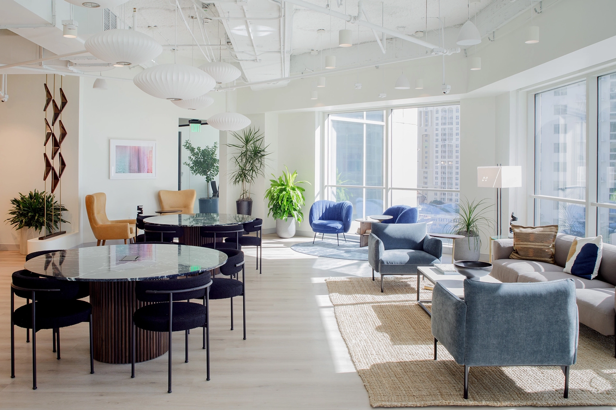 A modern, well-lit office lounge area in Fort Lauderdale features round tables with black chairs, blue armchairs, a gray sofa, and numerous indoor plants near large windows with city views—a perfect workspace or informal meeting room.