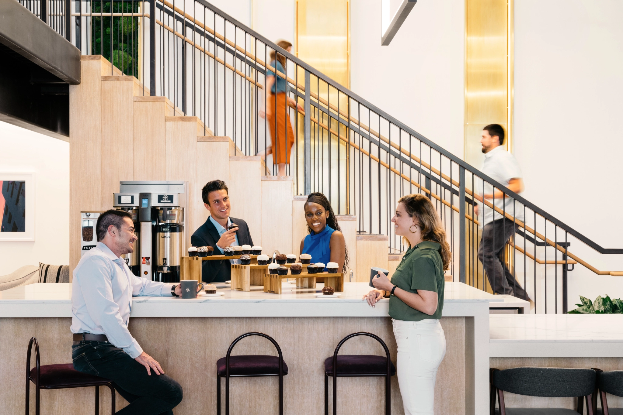 Vier collega's zitten rond een tafel met koffie en gebak in een gezellige werkruimte. Twee personen lopen op de achtergrond over de trap, wat bijdraagt aan de bruisende ambiance van het kantoor.