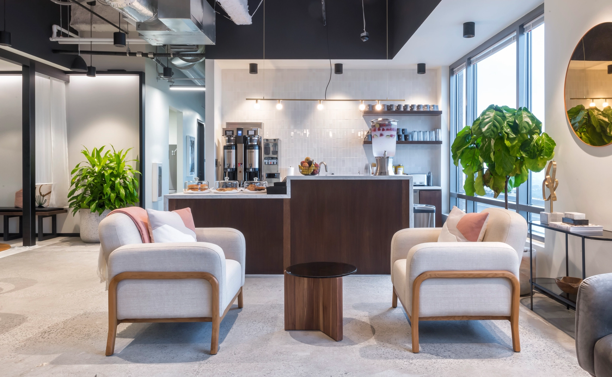 Moderne coworking-loungeruimte met twee gestoffeerde fauteuils, een kleine ronde tafel en een koffiestation op de achtergrond. Grote ramen en groene planten dragen bij aan de inrichting, waardoor het een uitnodigende ruimte is voor informele vergaderingen of ontspannen kantoorpauzes.