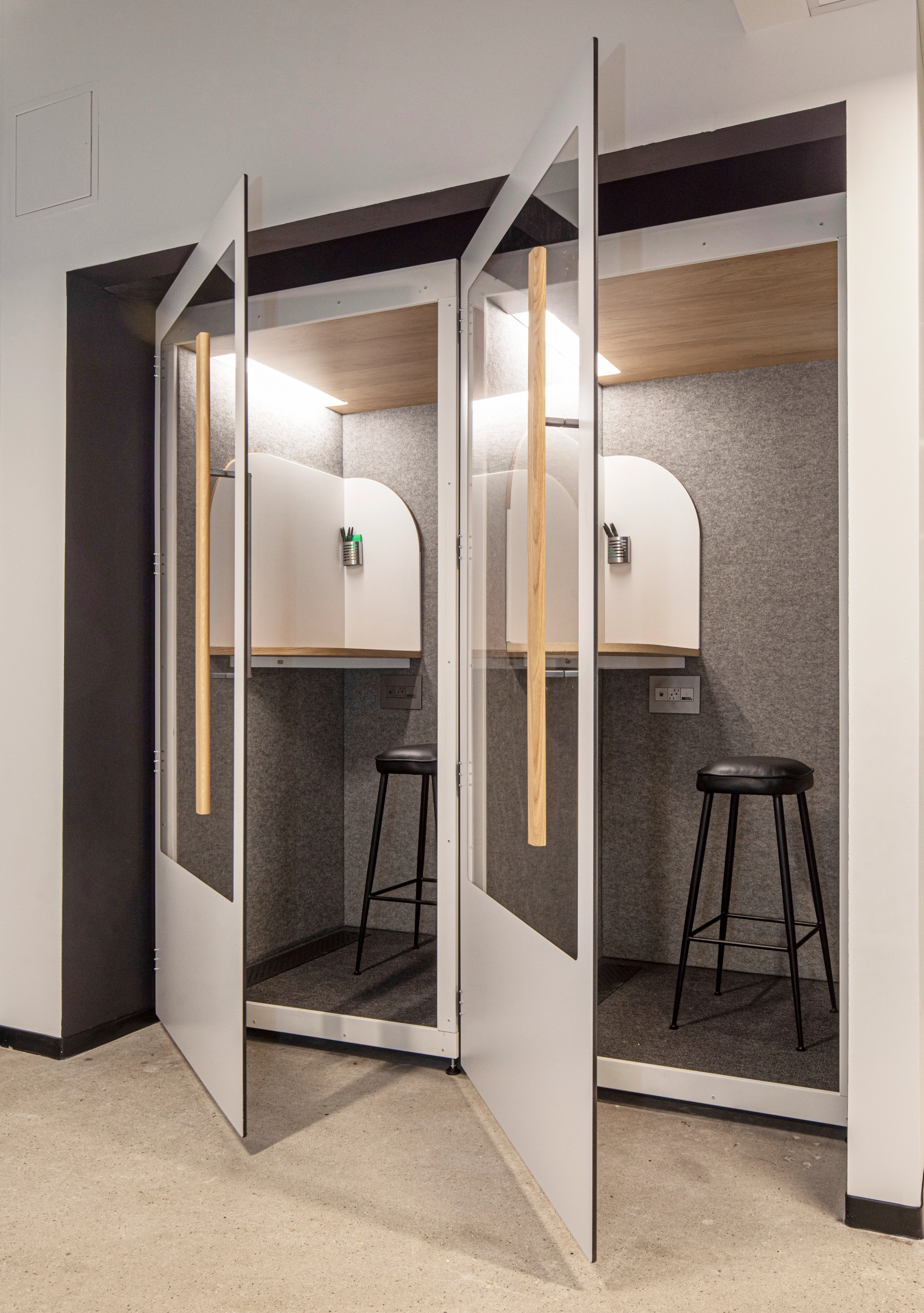 Two telephone booths with open doors reveal interiors with high stools and small desks with light grey walls, which fit perfectly into the collaborative atmosphere of the coworking space.