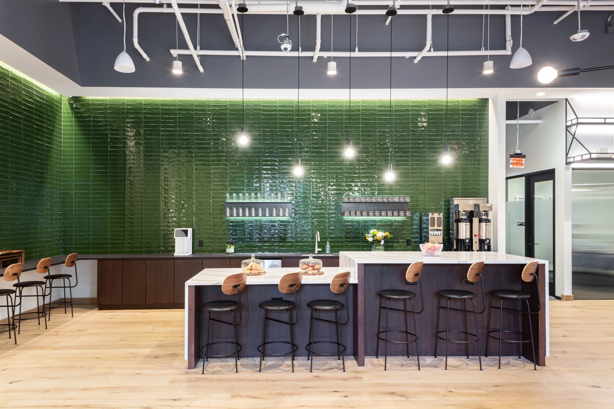 Een moderne keuken in een coworkingruimte in Dallas heeft een lange bar met hoge krukken, een groen betegelde muur en hanglampen.