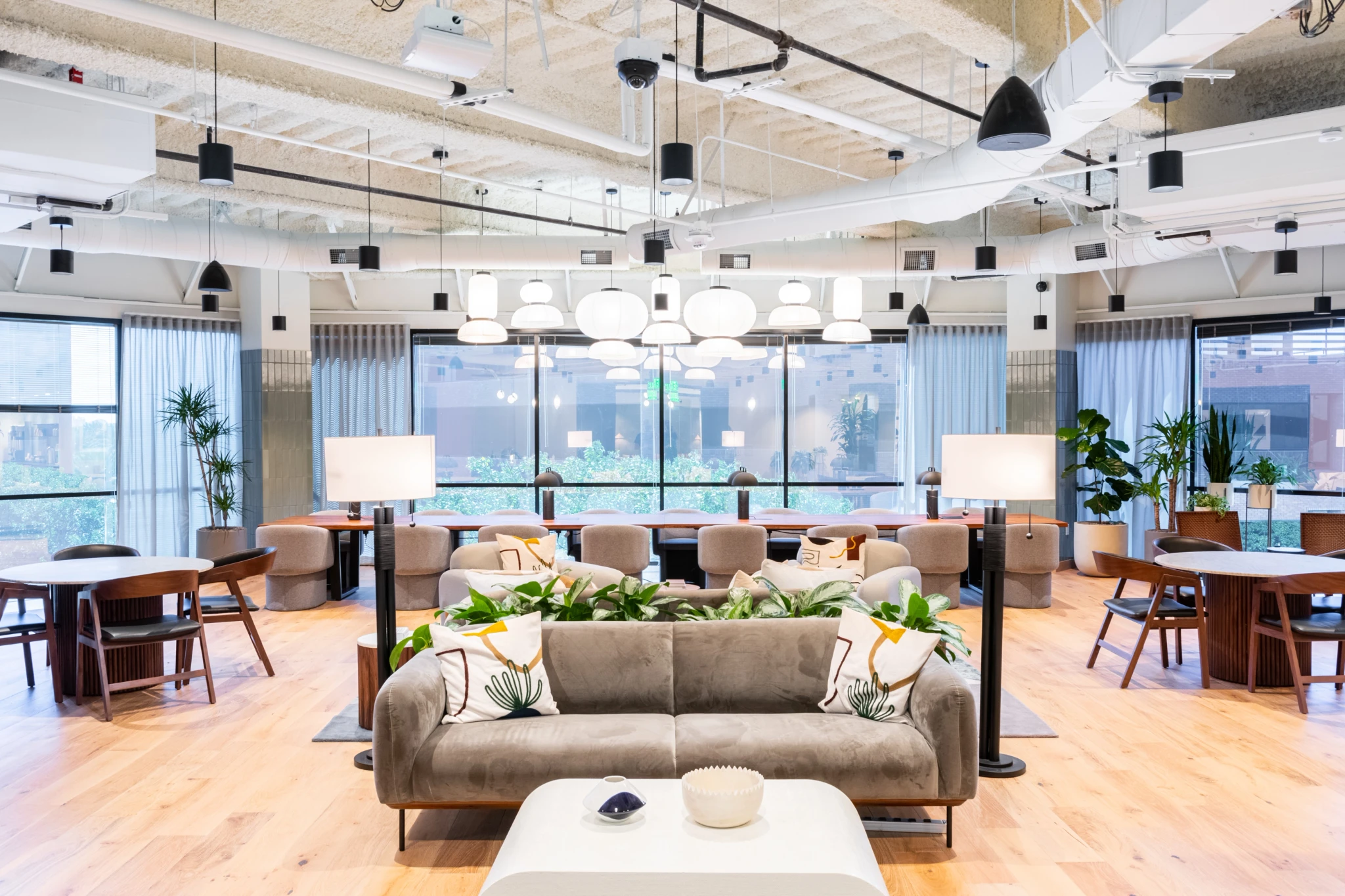 Een moderne, open coworkingruimte met houten vloeren, banken, tafels, planten en grote ramen. Heldere bovenverlichting en neutrale tinten creëren een ruime en uitnodigende sfeer, ideaal voor samenwerking of het houden van vergaderingen.