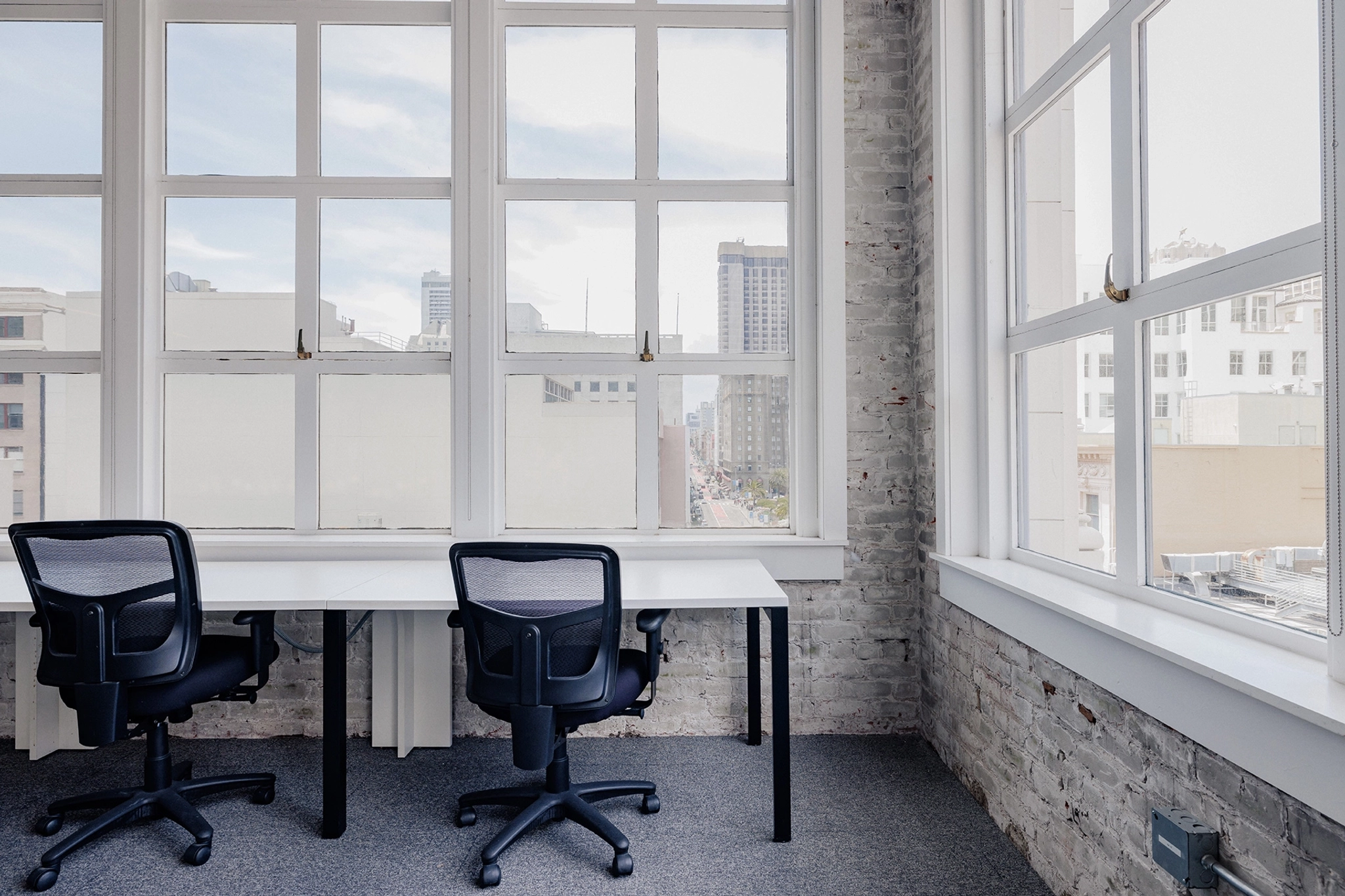 Dos sillas de oficina vacías se sientan frente a un escritorio blanco en un luminoso espacio de coworking, enmarcado por grandes ventanales con vista al ajetreado horizonte de San Francisco.