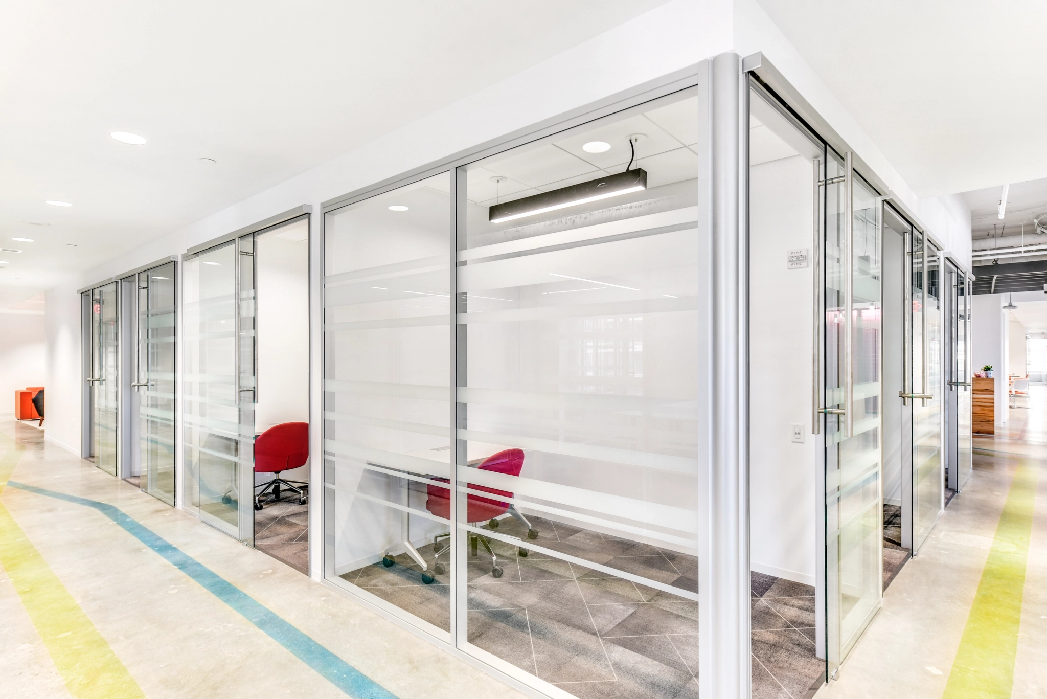 A modern coworking space in Arlington, featuring glass-enclosed meeting rooms with red chairs and clean, white walls. The hallway floor is adorned with multicolored lines.