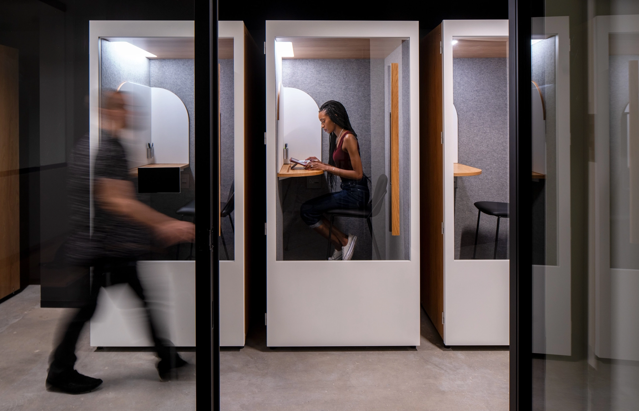 A man is walking past a women working in a phone booth in a coworking space.