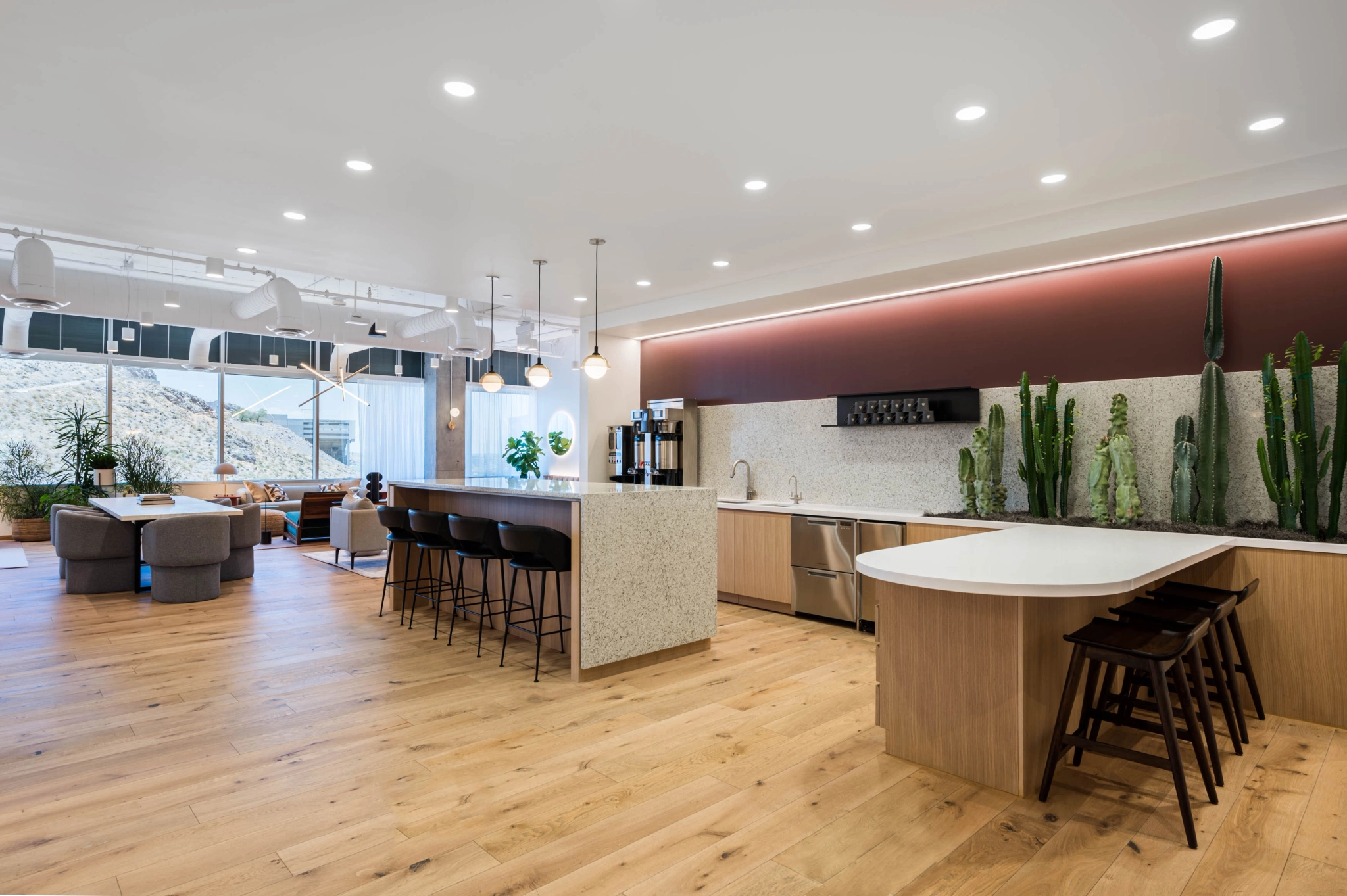 Moderna cocina abierta y sala de estar con pisos de madera, sillas de bar, encimeras blancas, lámparas colgantes y cactus altos en la pared; se puede utilizar a la perfección como un elegante espacio de coworking. Los grandes ventanales ofrecen vistas a la montaña, fomentando la creatividad y la productividad.