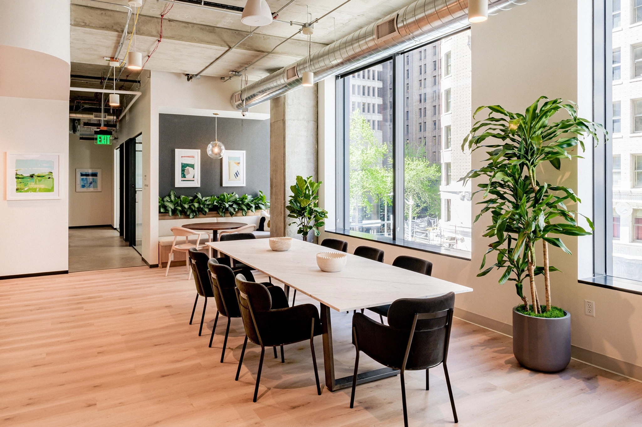 Un espacio de coworking moderno con una mesa larga, elegantes sillas negras y grandes ventanales. La animada oficina está decorada con varias plantas en macetas y decoraciones artísticas en las paredes, creando un lugar de trabajo inspirador para todos.