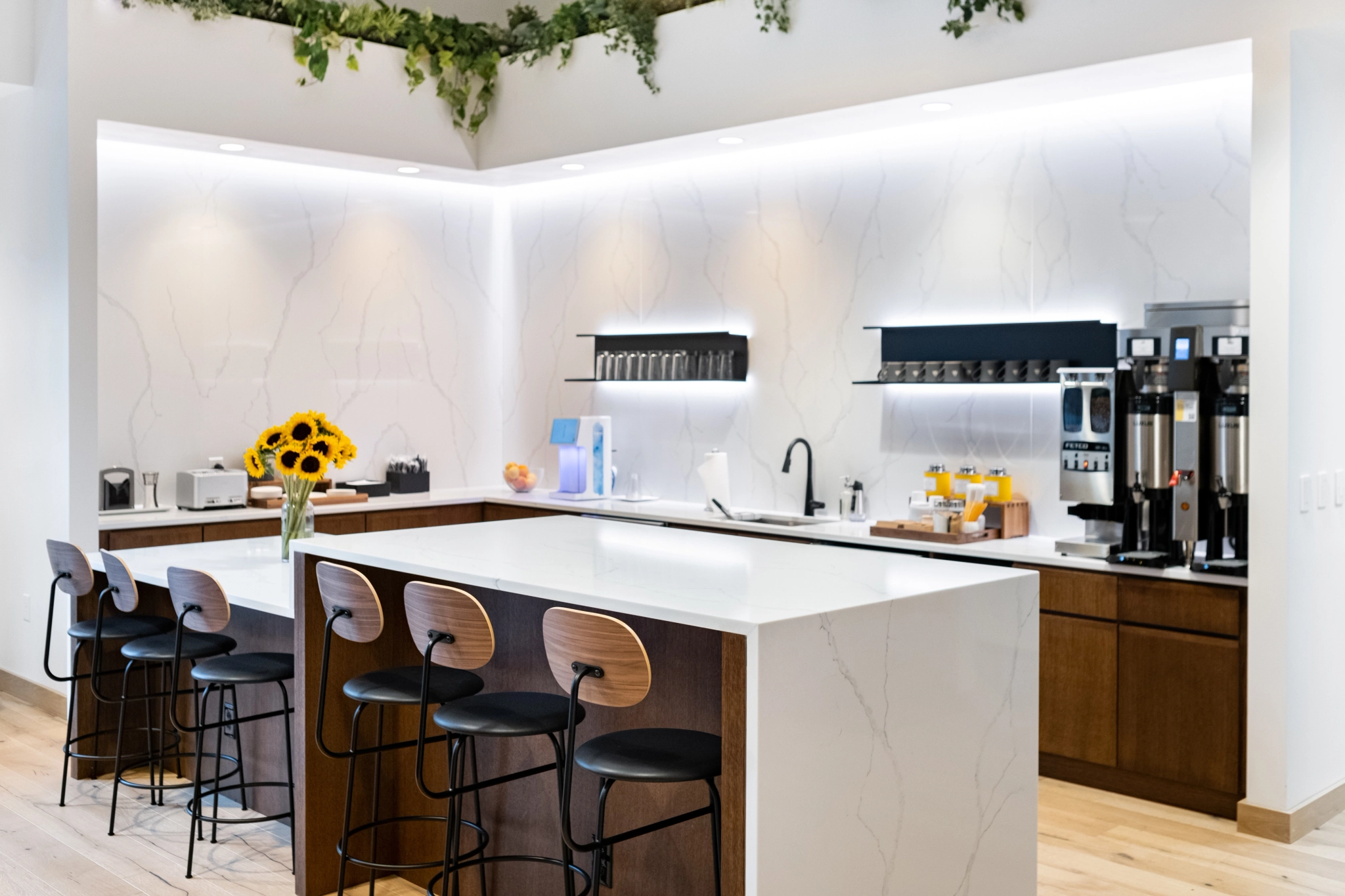 Moderne keuken met eiland, barkrukken en een vaas met zonnebloemen, die doet denken aan een trendy co-working space in Seattle. Witte, marmerachtige oppervlakken en koffiezetapparaten complementeren de planten die aan het plafond hangen.