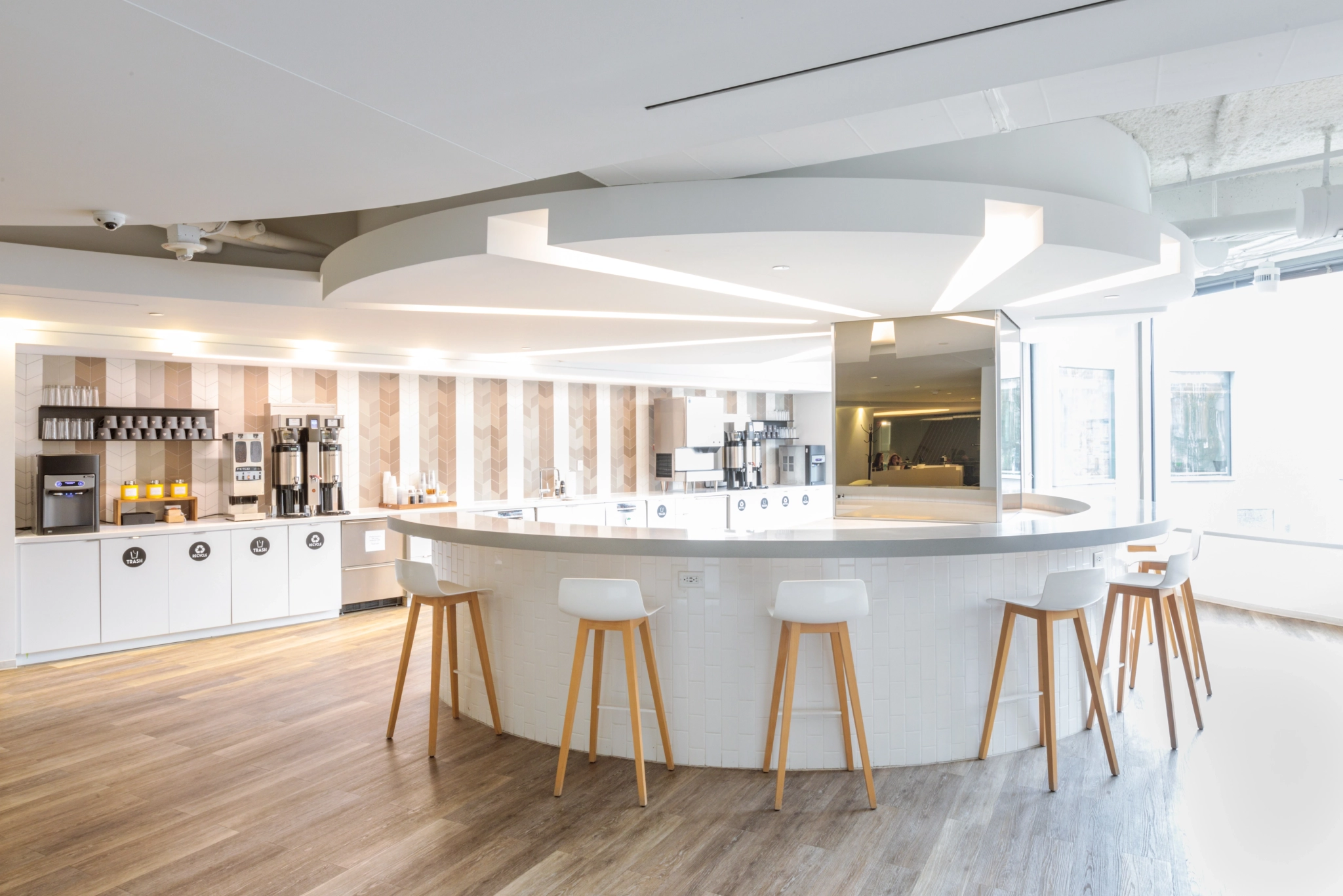 Moderne runde Kaffeebar mit weißen Arbeitsplatten, Holzhockern und mehreren Kaffeemaschinen vor einem Hintergrund in Weiß und hellem Holz, perfekt für einen Büroraum in Arlington.