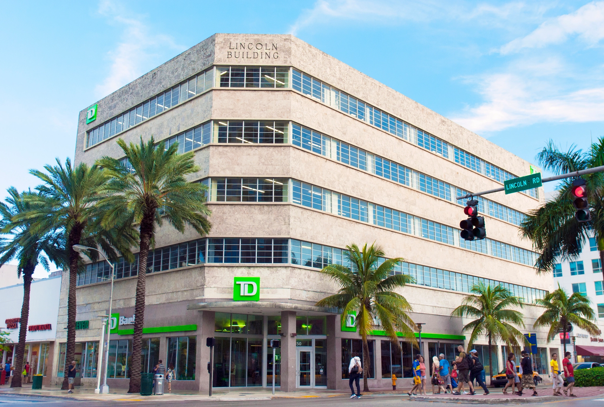 Un moderno edificio comercial de varios pisos en Miami Beach alberga un banco TD en la planta baja, rodeado de palmeras en una intersección de calles concurrida. Arriba, los espacios de coworking y las salas de reuniones ofrecen entornos de trabajo vibrantes en días claros y soleados.