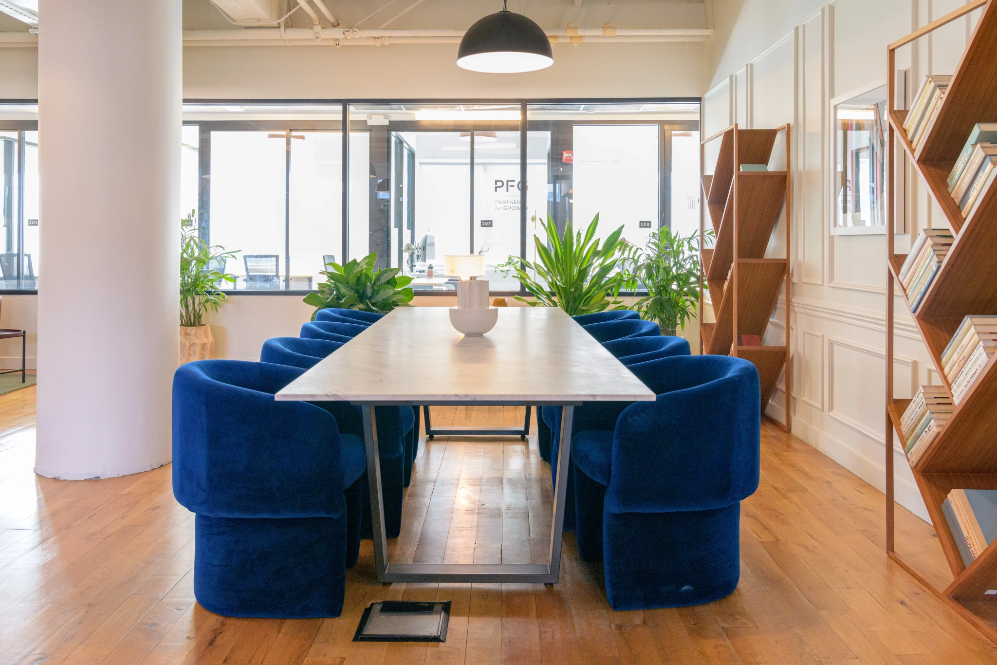 Een coworkingruimte met een vergaderruimte met een lange tafel, blauwe stoelen en glazen wanden. Planten en boekenplanken zorgen voor een vleugje groen, terwijl een witte kom elegant op tafel staat.