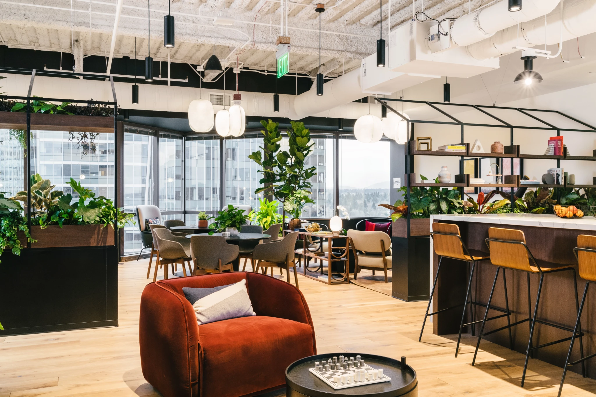 Een moderne coworking lounge met kamerplanten, ronde tafels, stoelen en een bruine fauteuil. Planken en barkrukken bevinden zich op de achtergrond, onder hangende lampen en zichtbare plafondkanalen, wat een uitnodigende werksfeer creëert.